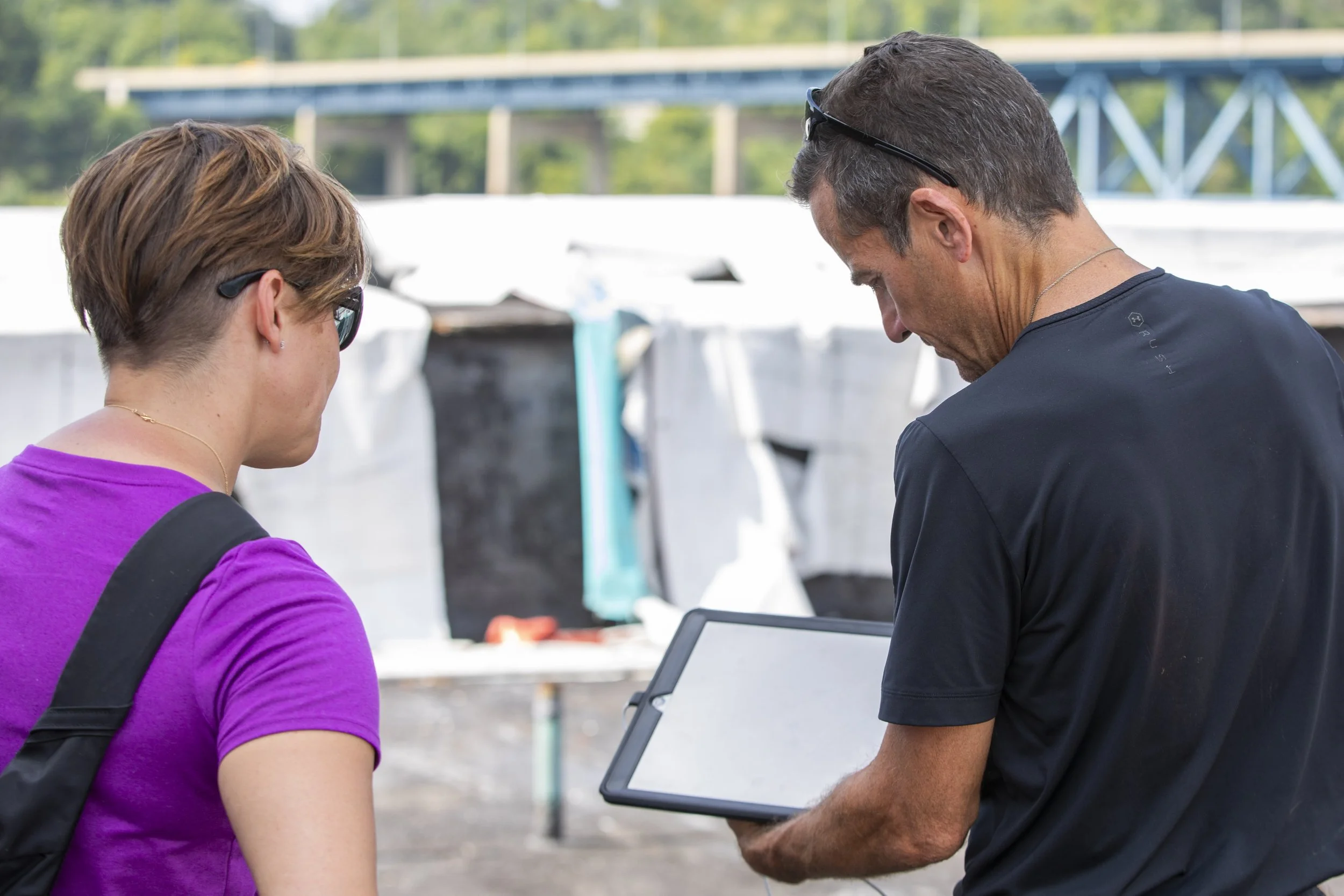 Two people, one with short brown hair in sunglasses wearing a purple shirt and the other with gray hair in a black shirt and glasses on his head, are looking at a tablet outdoors near water with boats and a bridge in the background.