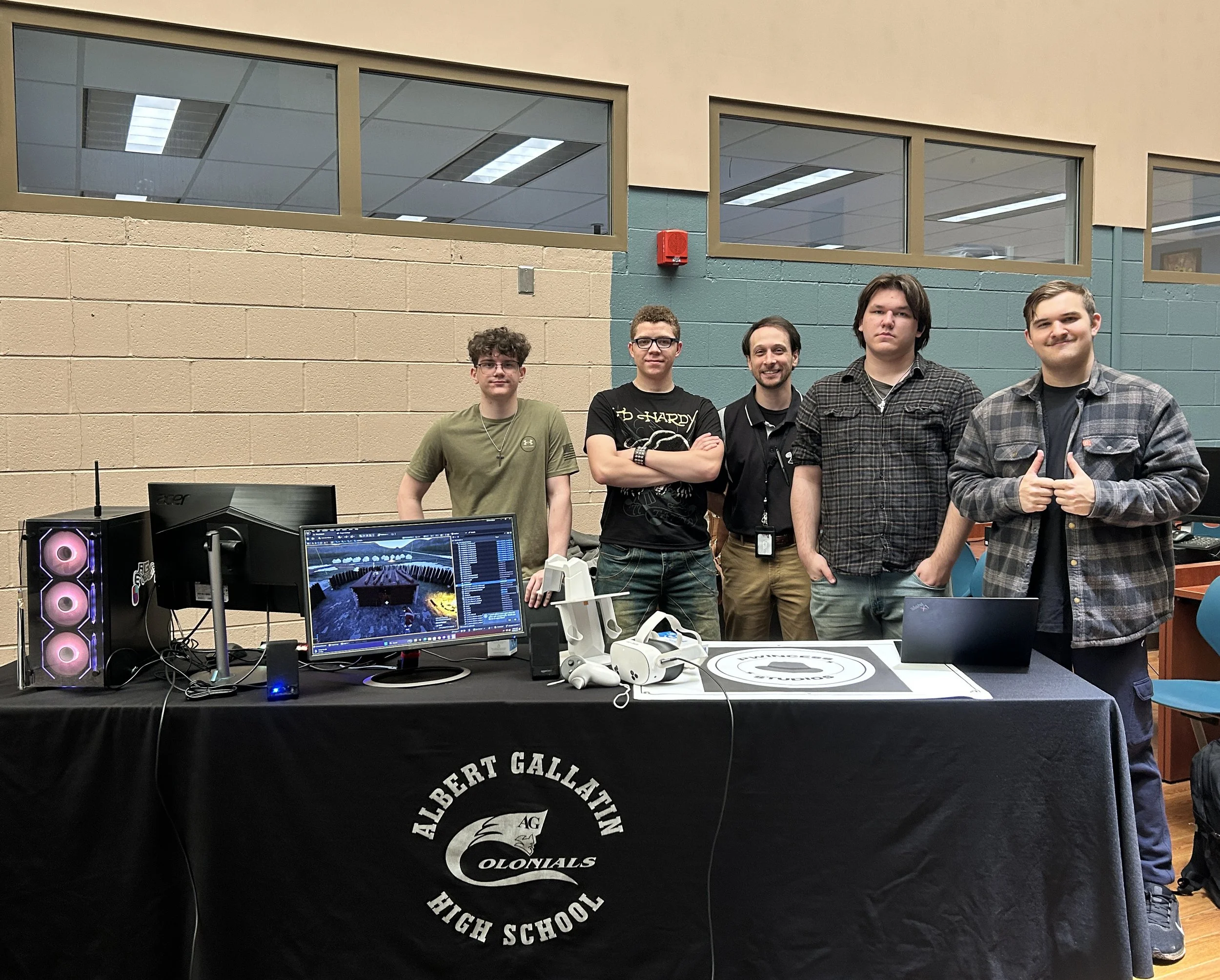 A group of five young men standing behind a table with gaming and computer equipment at Albert Gallatin High School. The table is covered with a black cloth with the school's name and logo. The background shows windows and a beige brick wall.