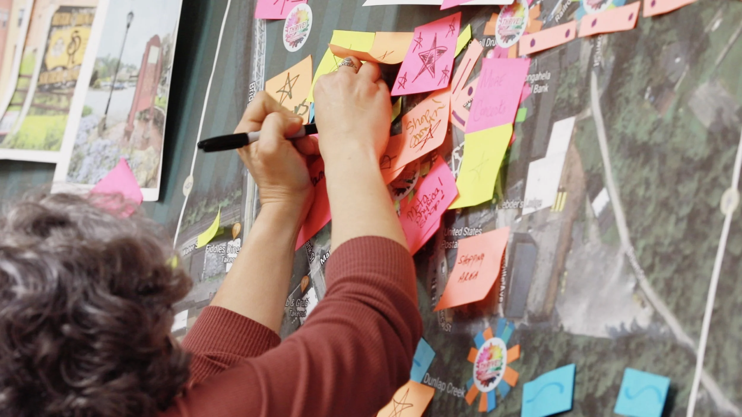 A person writes on a pink sticky note with a black marker, surrounded by other colorful sticky notes on a wall or table, with pictures and signs in the background.