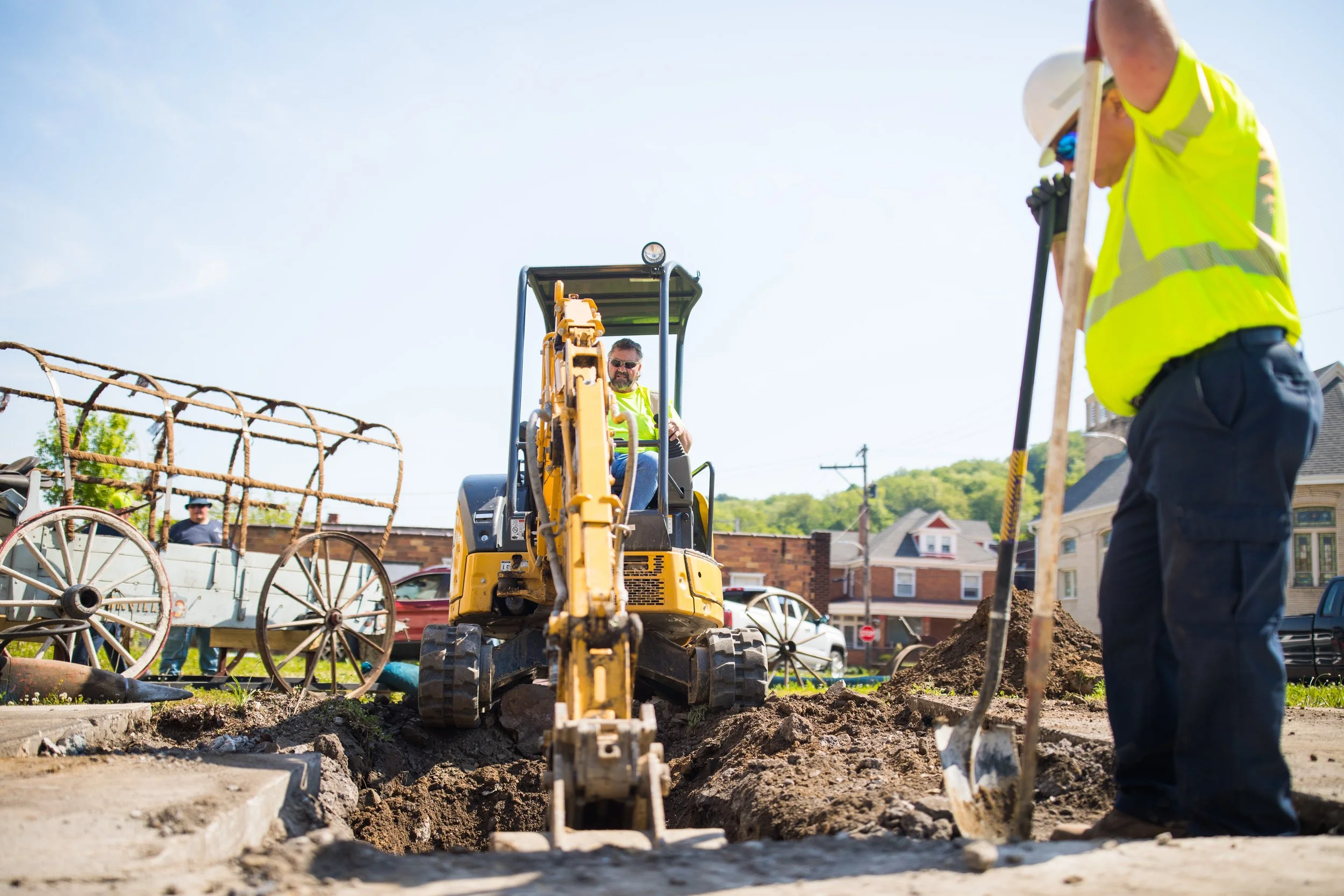 Construction workers are working on a sidewalk project using heavy machinery and shovels in a neighborhood with houses and green hills in the background.