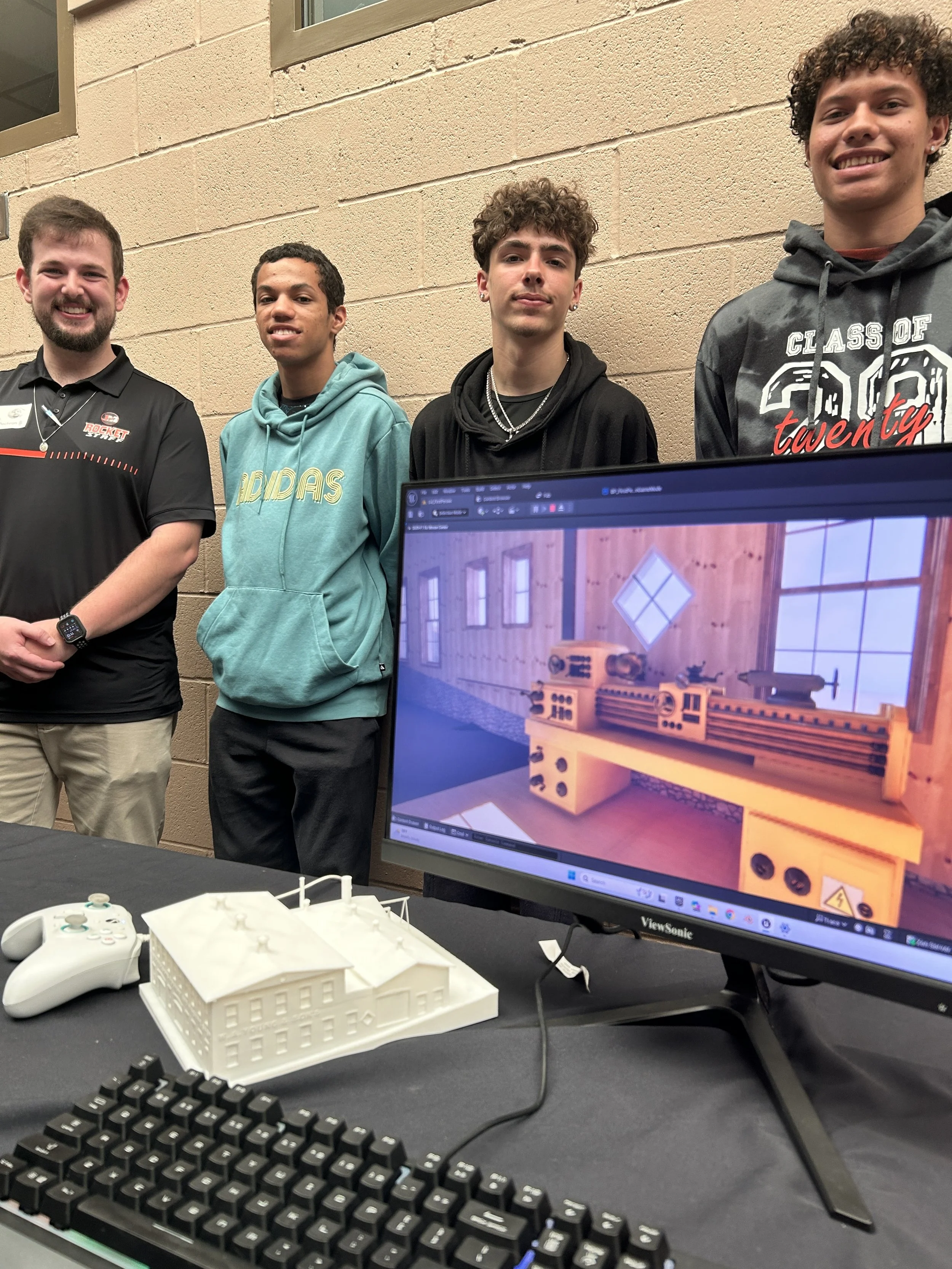Four young men standing behind a table with a white architectural model, a computer monitor displaying a 3D design of a wooden structure, and gaming controllers in the foreground.