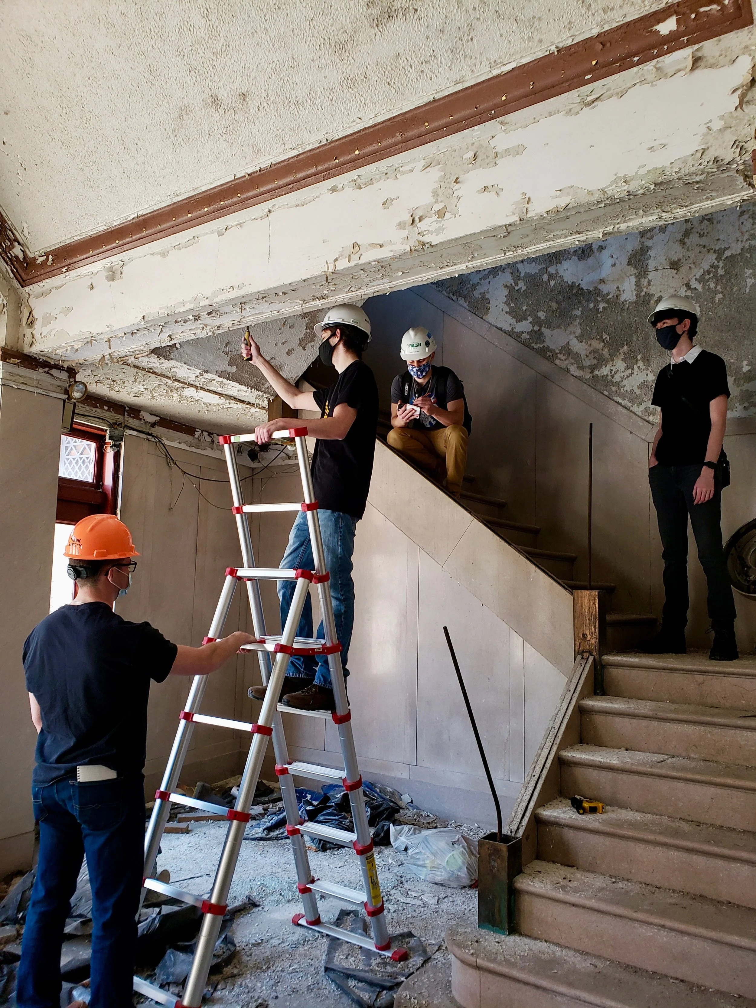 Four workers in hard hats and masks working on renovation of a building's interior. One worker is on a ladder, others are on the ground near the staircase, which is under construction or demolition.