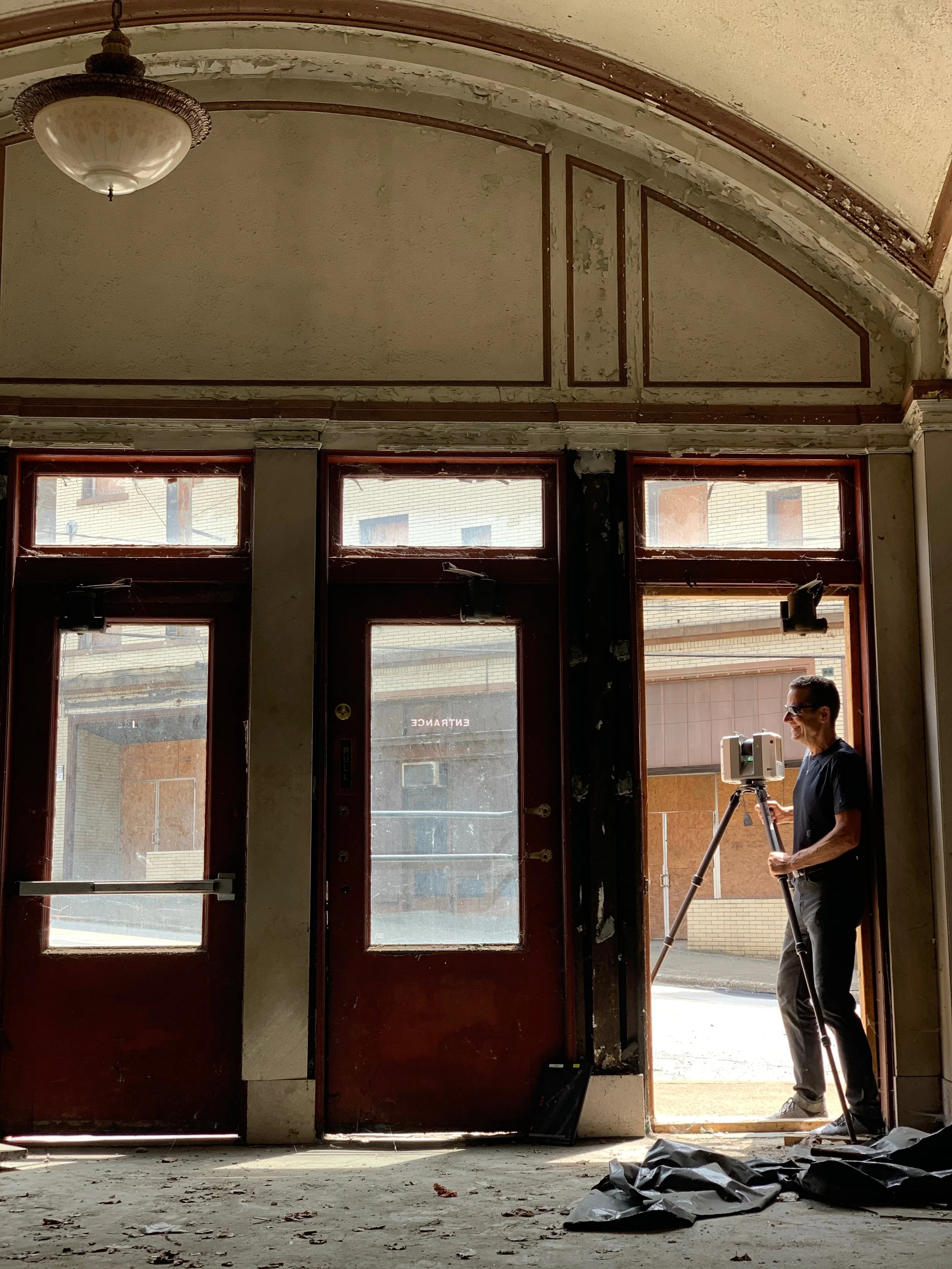 A person wearing glasses and a black shirt is standing on a tripod inside a building with peeling paint and broken windows, near an open door with sunlight coming through.