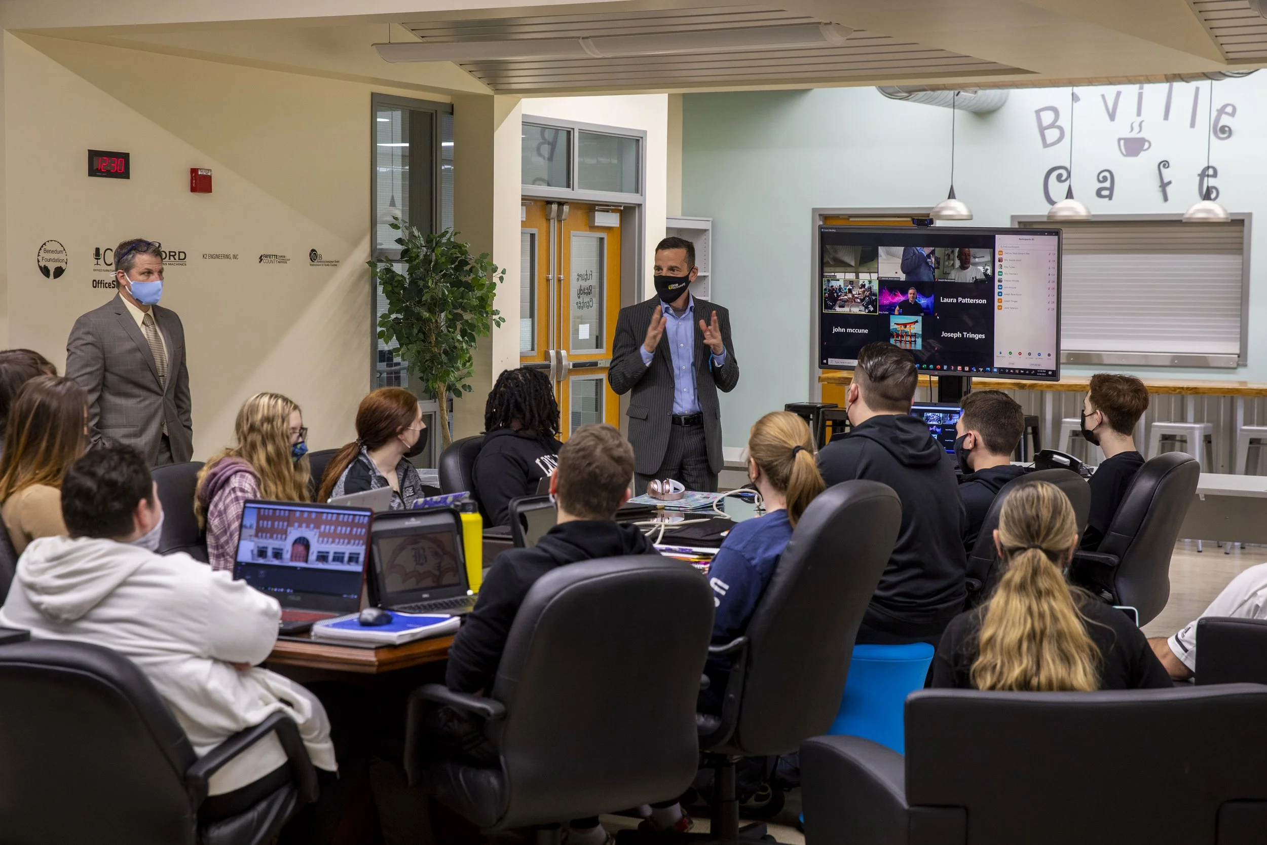 A classroom or conference room filled with students or attendees listening to a man in a pinstripe suit and black mask speaking. Other individuals, including a man in a gray suit, are present, all wearing masks. A large screen displays virtual meeting participants.