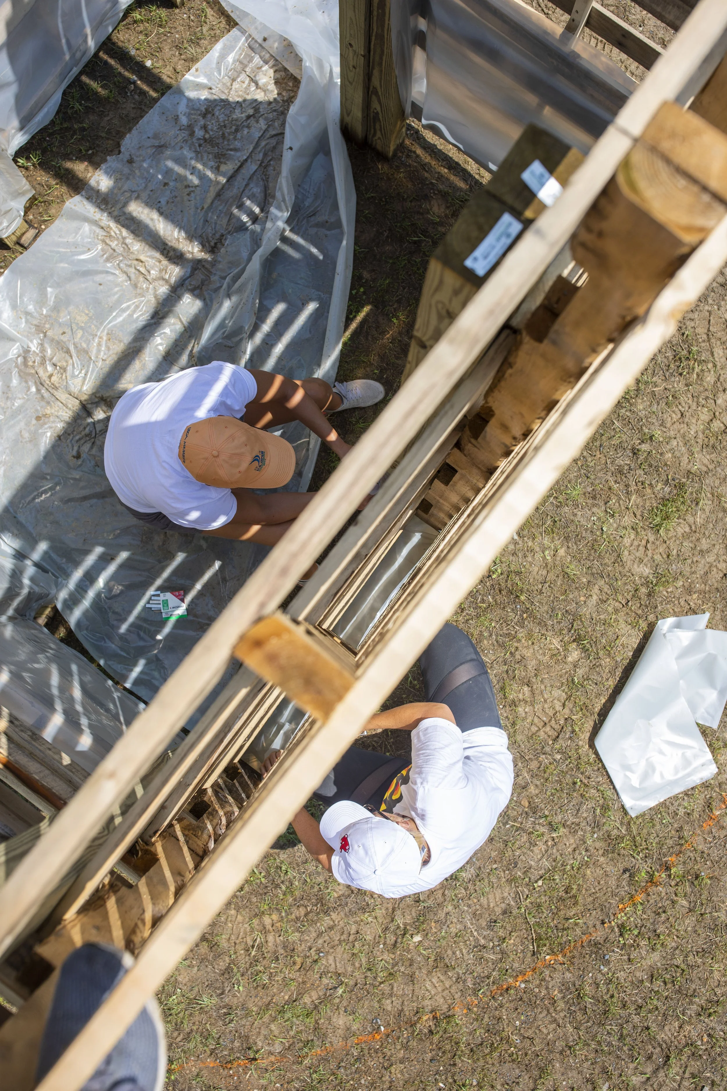 High-angle view of two people working on a construction site. One is wearing a white shirt and a beige cap, and the other is wearing a white shirt and has a white hard hat nearby. They are inside a partially built wooden structure with fencing and construction materials around them on the ground.