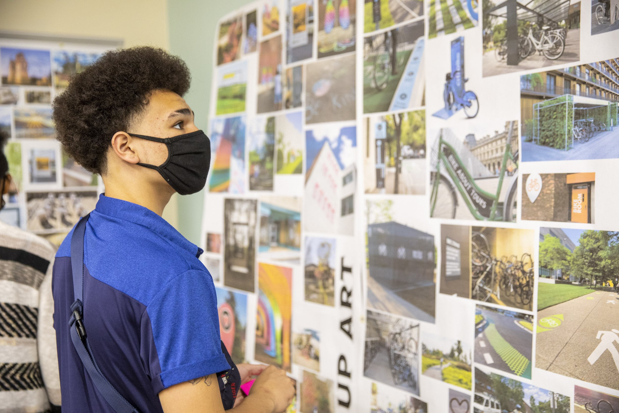 A young person with curly hair wearing a black face mask and a blue shirt looking at a display of colorful outdoor and biking photos on a wall.