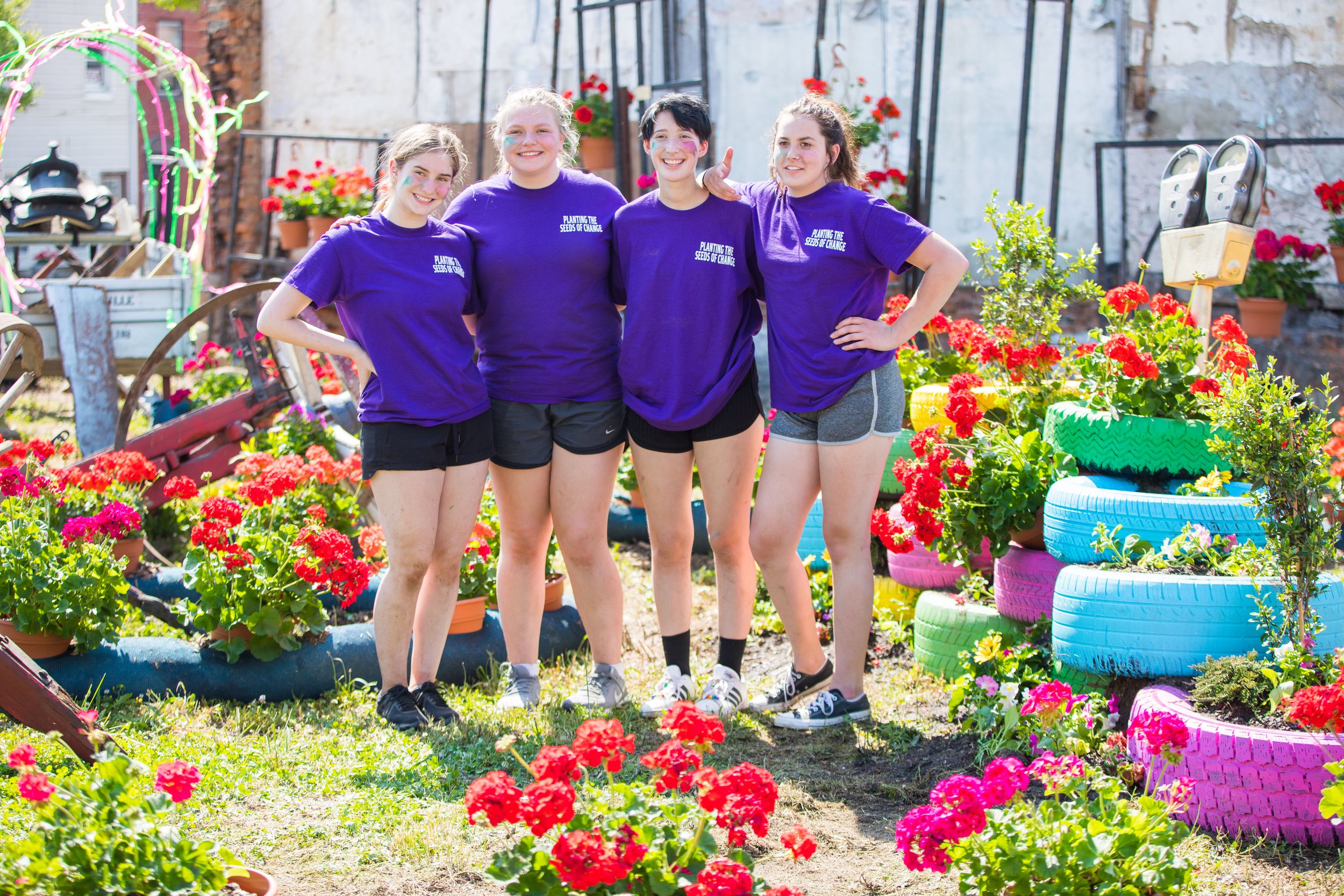 Four teenage girls wearing purple T-shirts with the text 'Planting the Seeds of Change' pose together in a flower garden, surrounded by colorful tires turned into flower beds and potted plants.