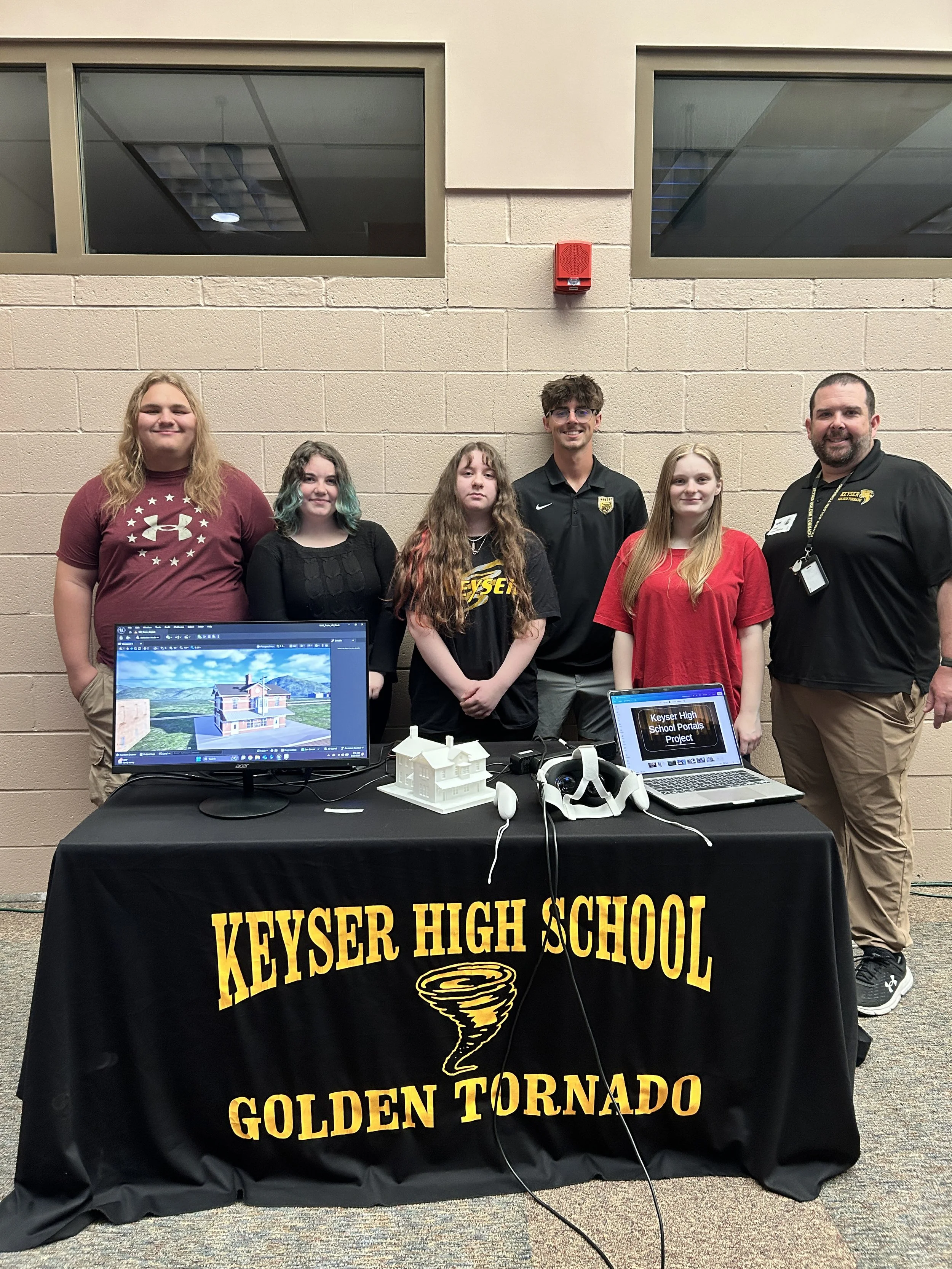 Group of six students and a teacher standing behind a display table with a black cover that has the words 'Keyser High School Golden Tornado' printed in yellow and a tornado illustration. There are desks with computer monitors in front of the table, one showing a 3D house model and another displaying 'Keyser High School Portals Project.' The students are smiling, and the setting appears to be a school event or presentation in a room with beige brick walls and high windows.