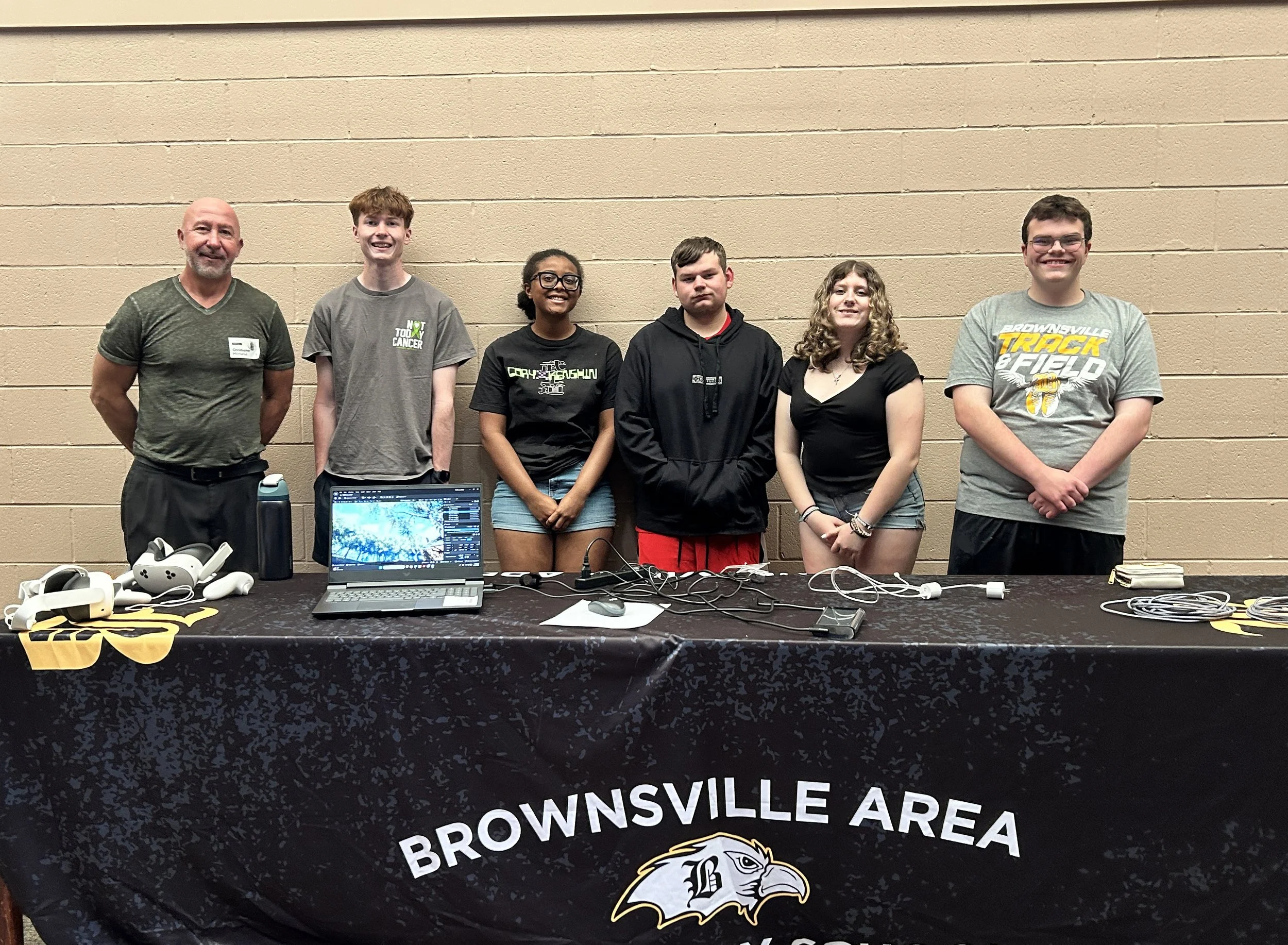Group of six diverse people standing behind a table with equipment, in front of a beige wall. The table has a computer, white headsets, and other electronic devices, with a black and yellow tablecloth bearing the words 'BROWNSVILLE AREA.'