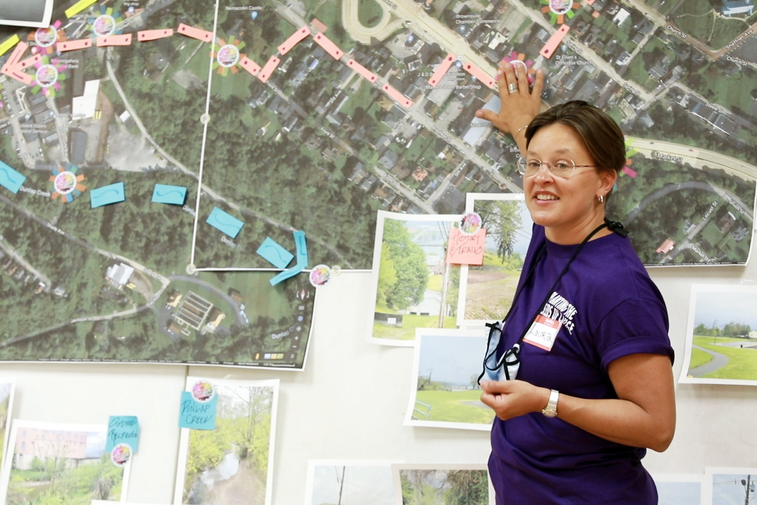 A woman in glasses and a purple shirt standing in front of a large map with various colorful markers, pointing at it with her right hand and smiling.