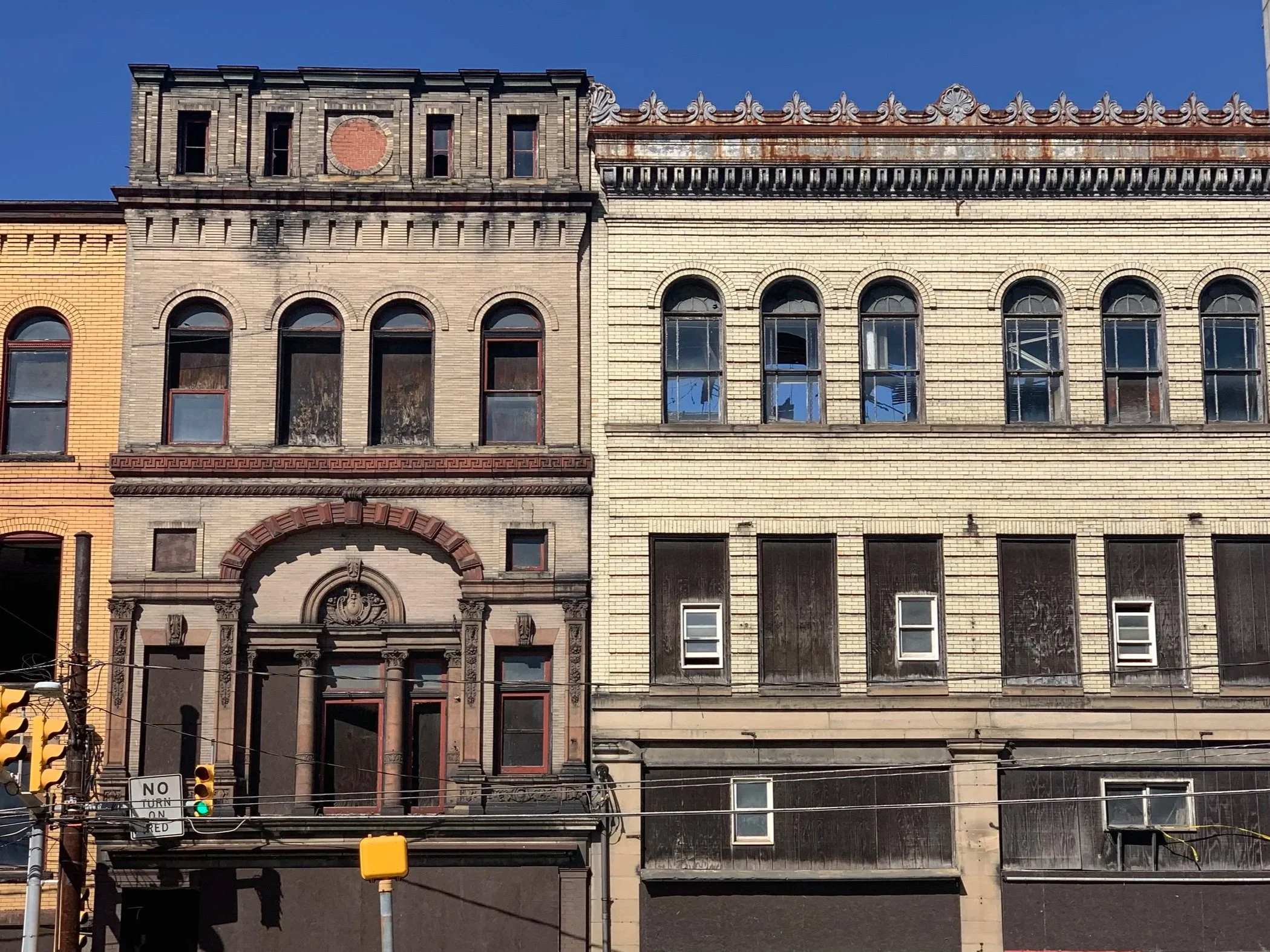 A row of old brick buildings with various architectural details, including arched windows, decorative cornices, and a mix of colors from yellow to beige and gray. There are utility wires and traffic signals in the foreground.