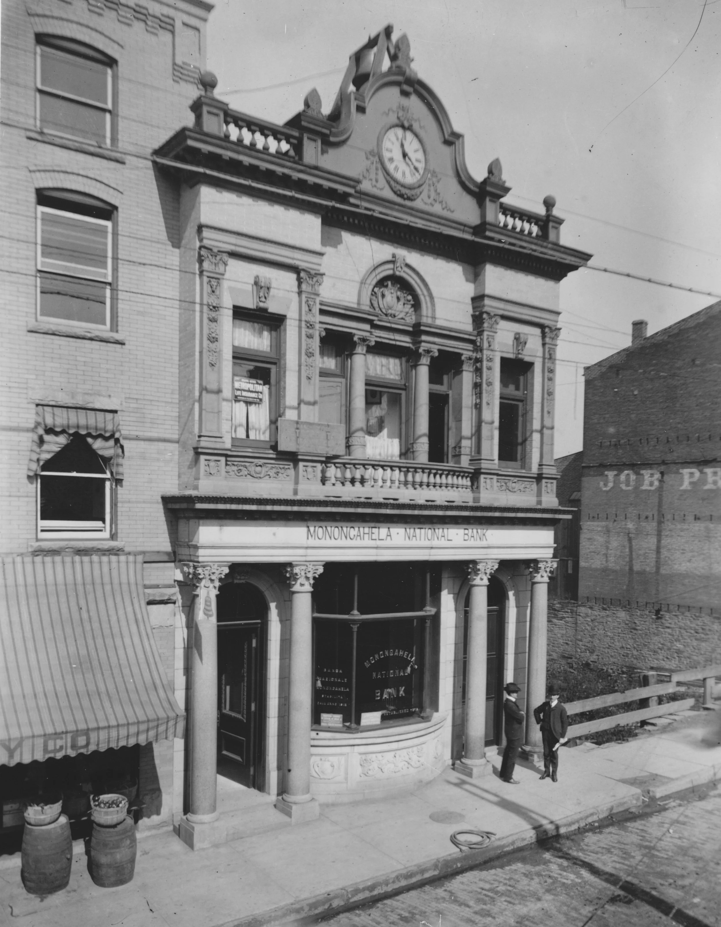 Black and white photo of a historic building with a clock on top, labeled 'Monongahela National Bank'. The building features ornate architectural details, columns, and large windows. Two men in suits stand outside, near a sidewalk with some barrels and a coiled hose.