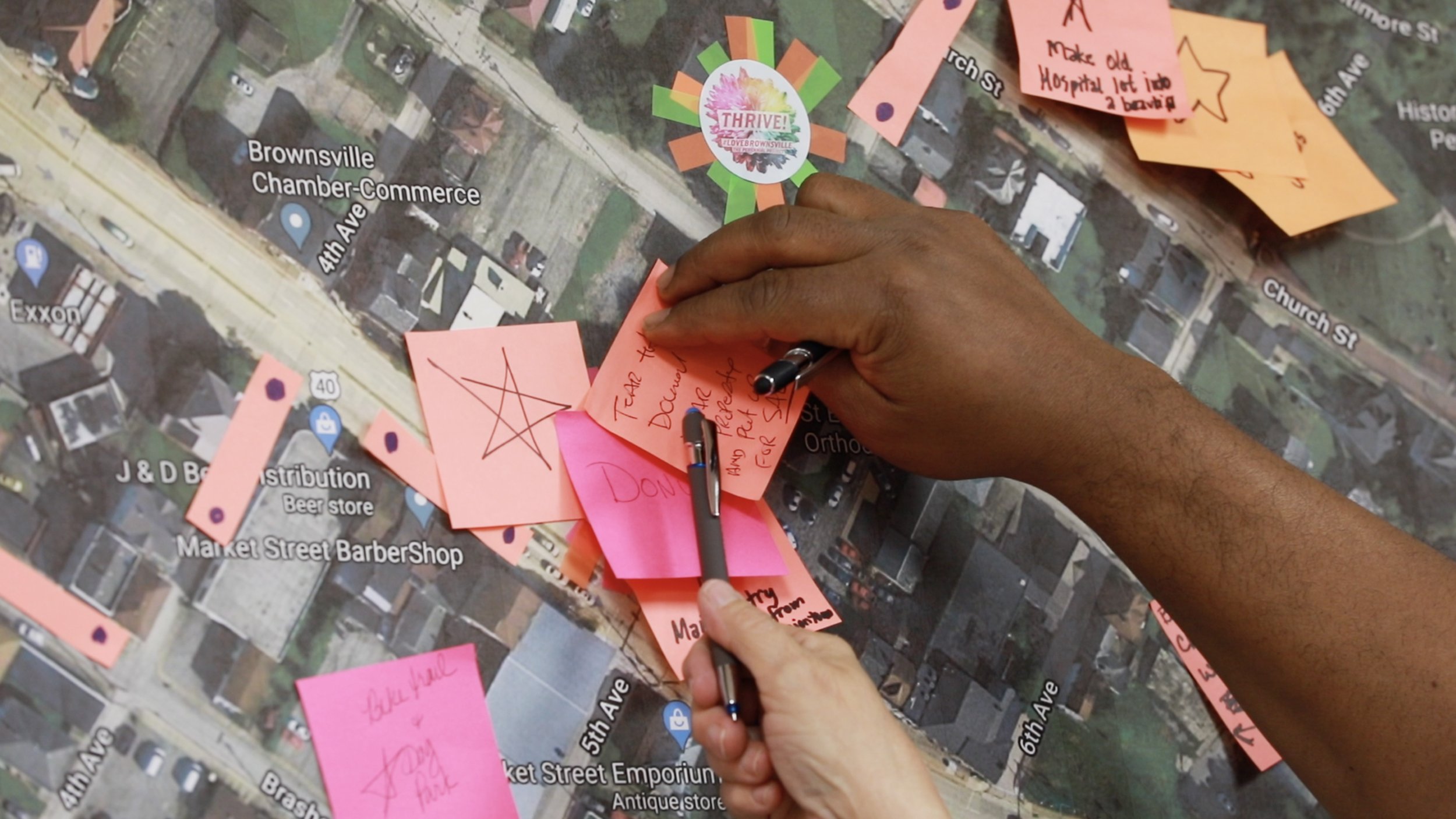 Hands writing on pink and orange sticky notes placed on a digital map of Brownsville, Tennessee, with streets and landmarks visible.