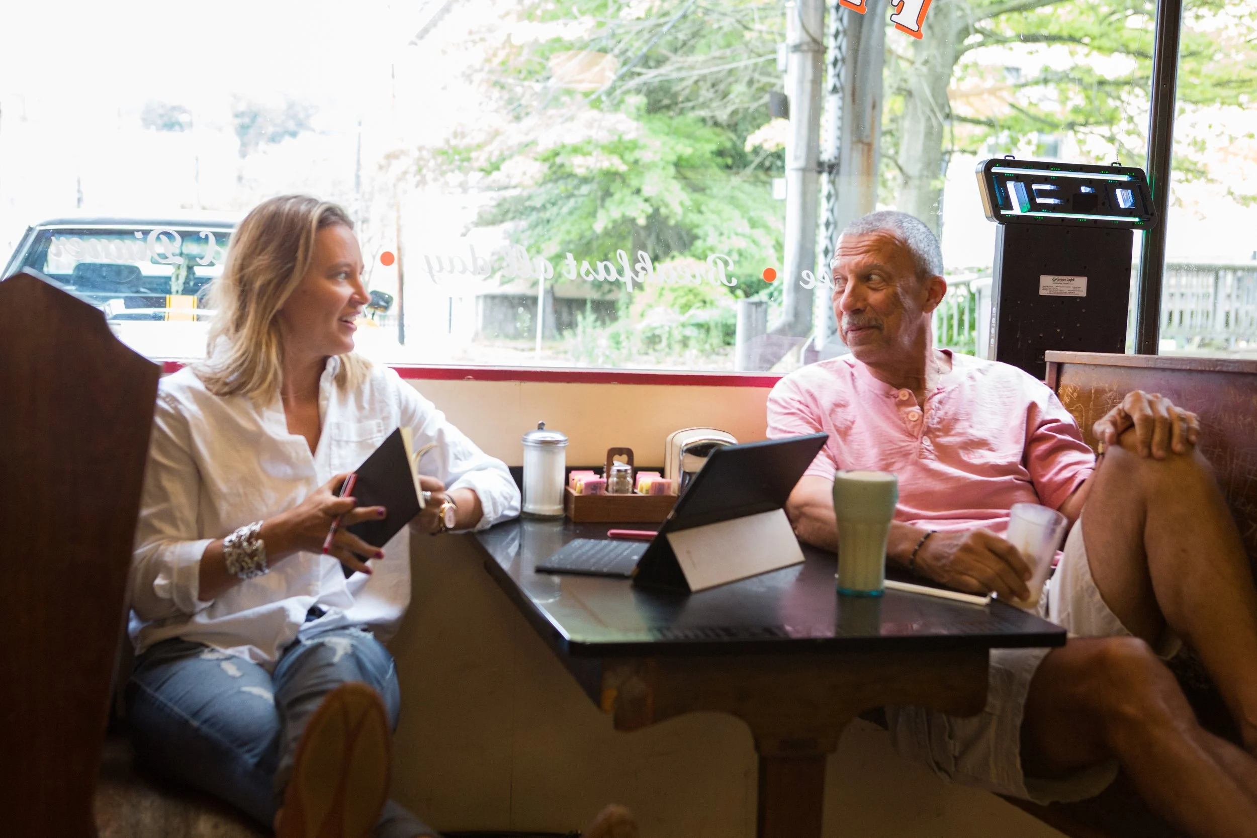 Four people sitting at a table inside a restaurant, engaged in conversation, with laptops and drinks on the table. Large windows reveal trees and power lines outside.