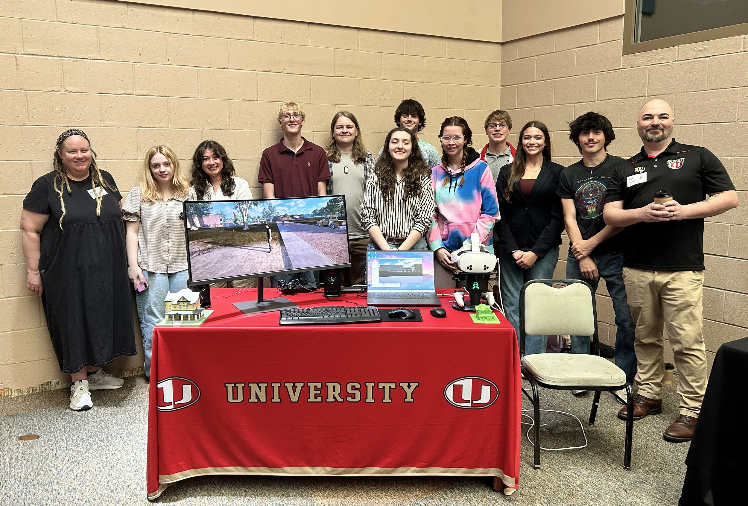 Group of diverse young adults and an older man standing behind a table with laptops, monitors displaying outdoor scenes, and VR equipment, at a university event with a red tablecloth featuring the university logo.
