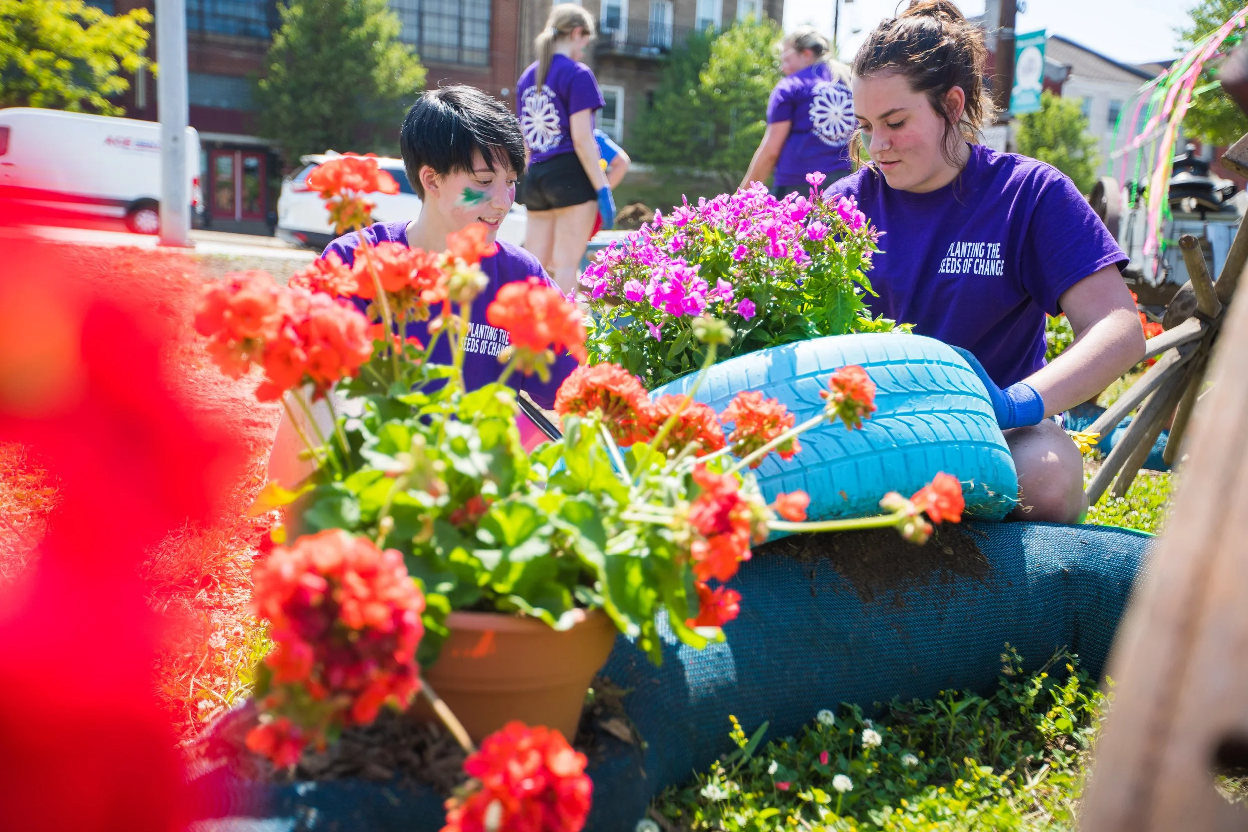 Two young women working together planting flowers in a garden bed outdoors on a sunny day, with additional people and buildings visible in the background.
