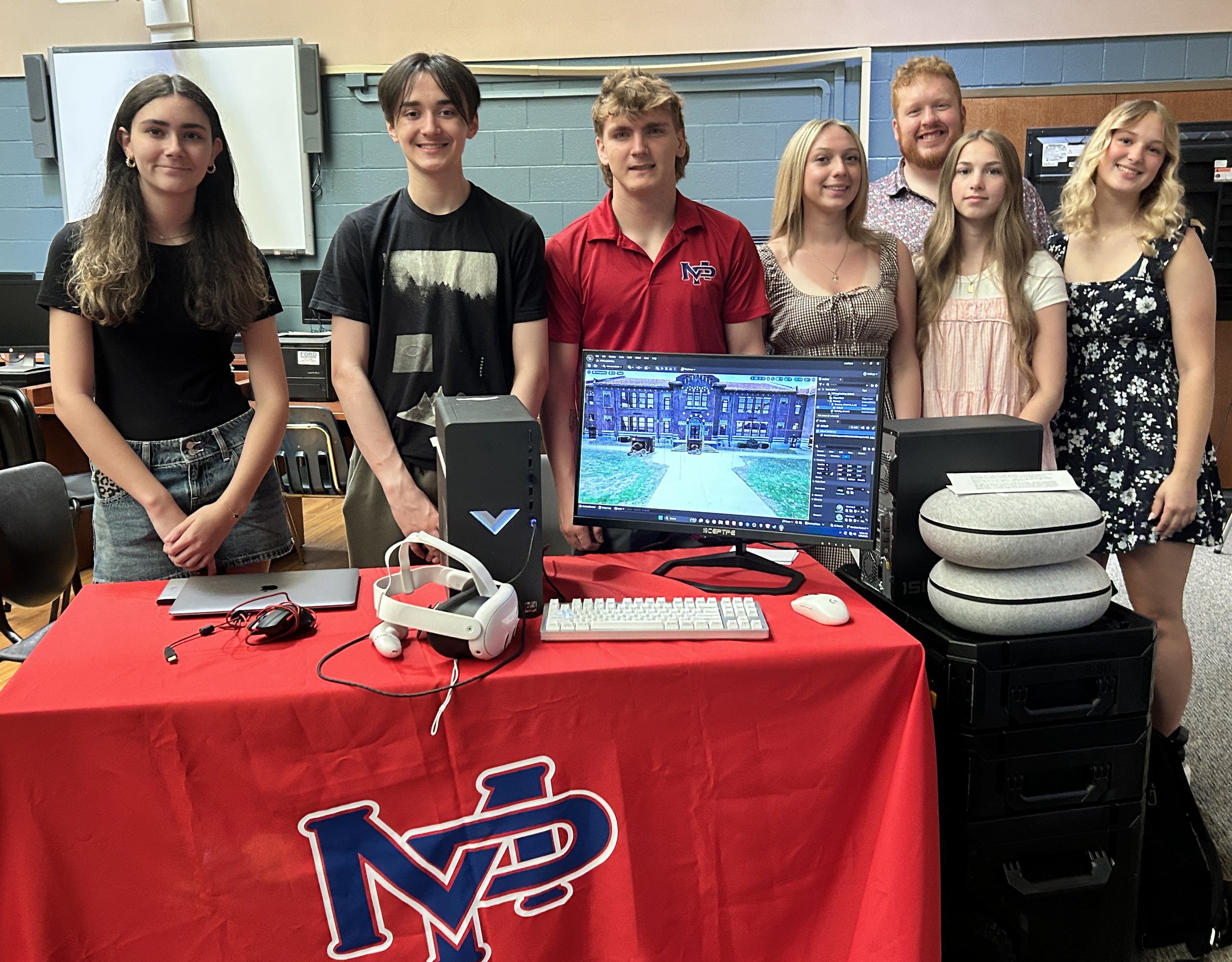 Group of seven young people standing behind a table at a tech or gaming event, with computer monitors, keyboards, headphones, and a desktop computer in front of them.
