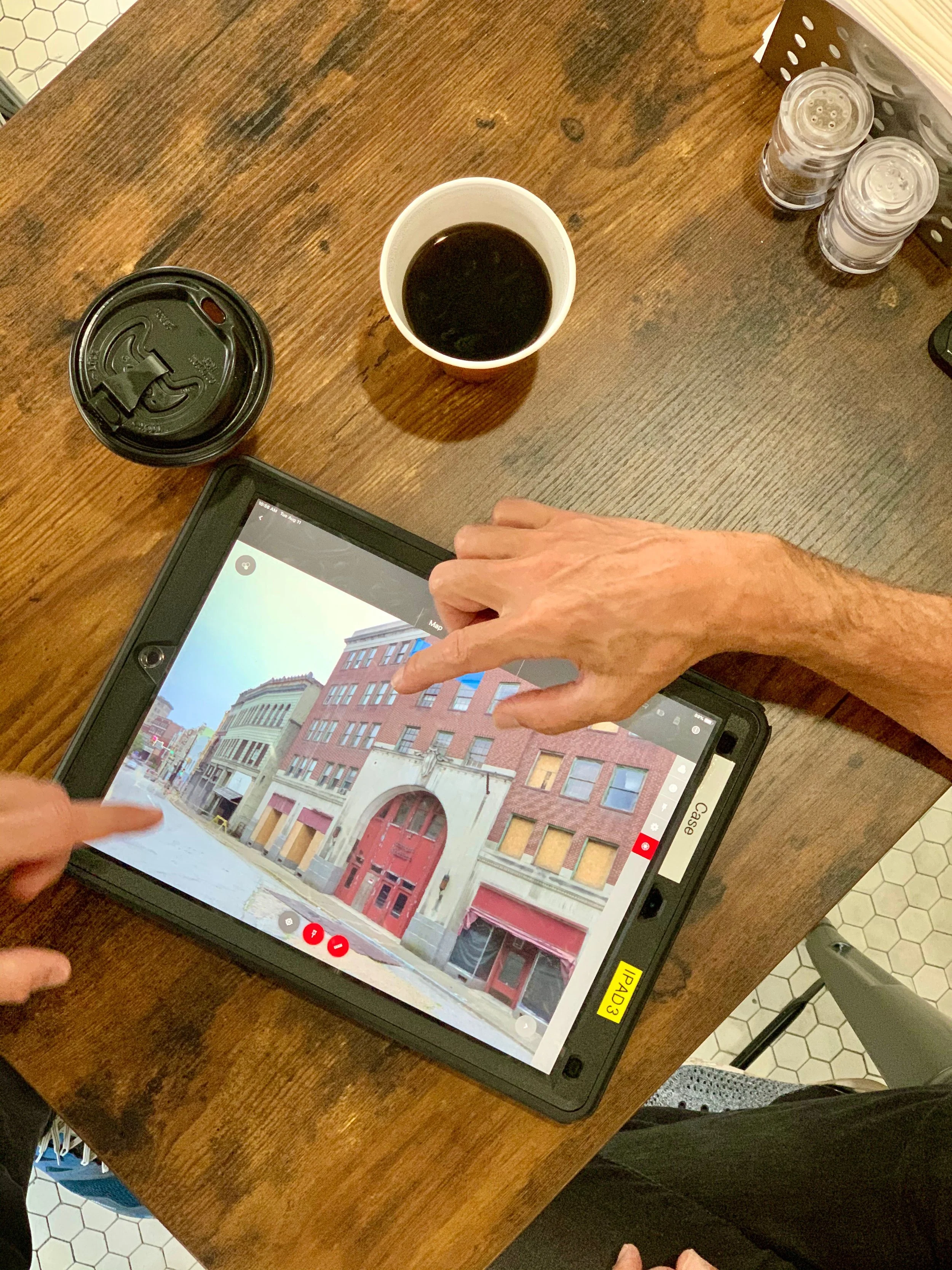 A person's hands pointing at a street view of buildings on a tablet at a wooden table. On the table, there are a black coffee cup, a takeout coffee cup with a black lid, and three small salt shakers. The tablet screen displays a street with buildings.