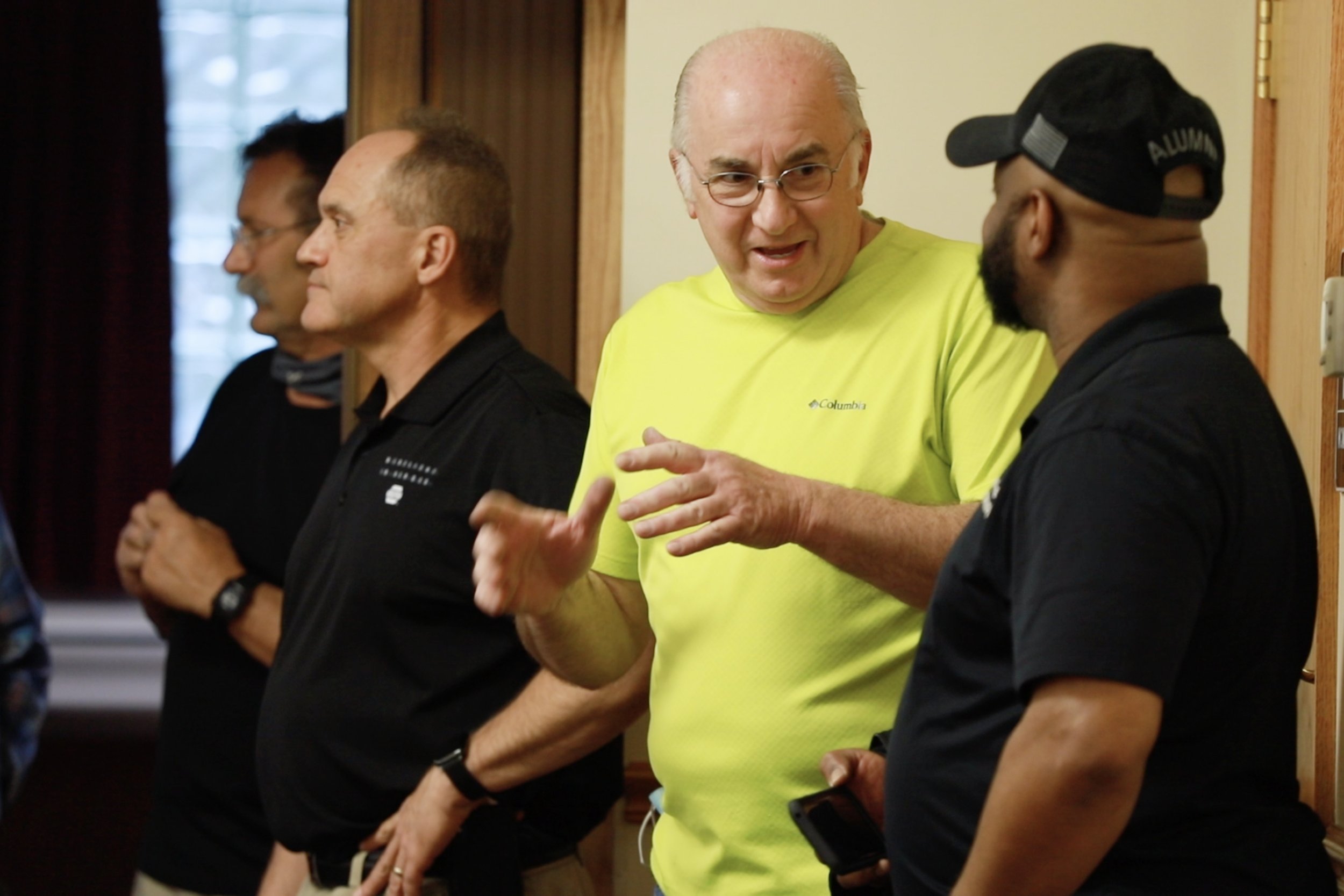 Four men standing in a line indoors, engaged in conversation. The man in the yellow shirt is speaking and gesturing, while the others listen. The men are dressed casually, and one is wearing a black cap and shirt. The background has wooden trim and a window.