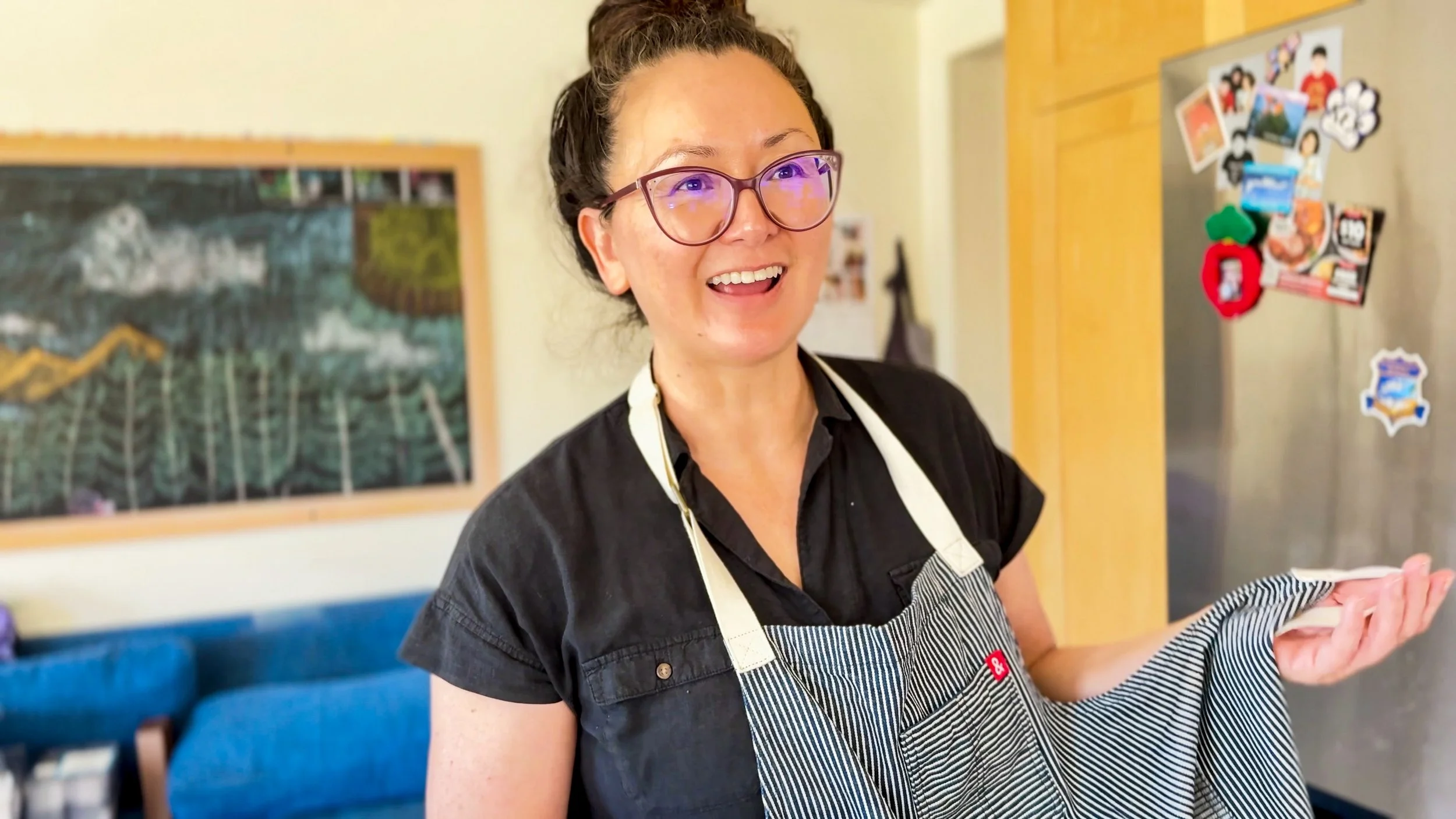 A woman with glasses and a bun hairstyle, smiling while wearing a black shirt and striped apron, possibly in a kitchen or restaurant setting.