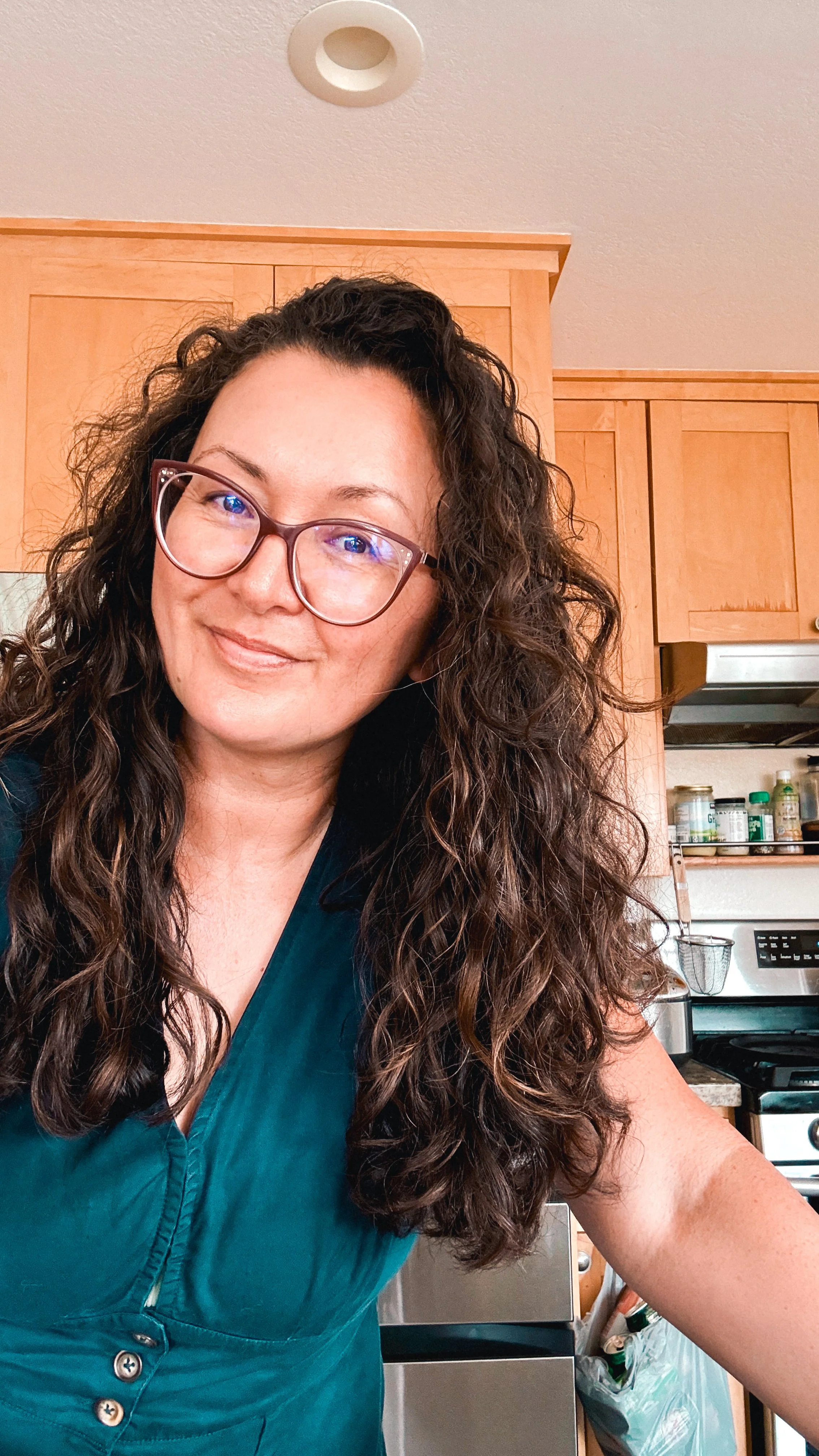 A woman with curly brown hair and glasses smiling in a kitchen, wearing a teal top, with wooden cabinets, stove, and kitchen supplies in the background.