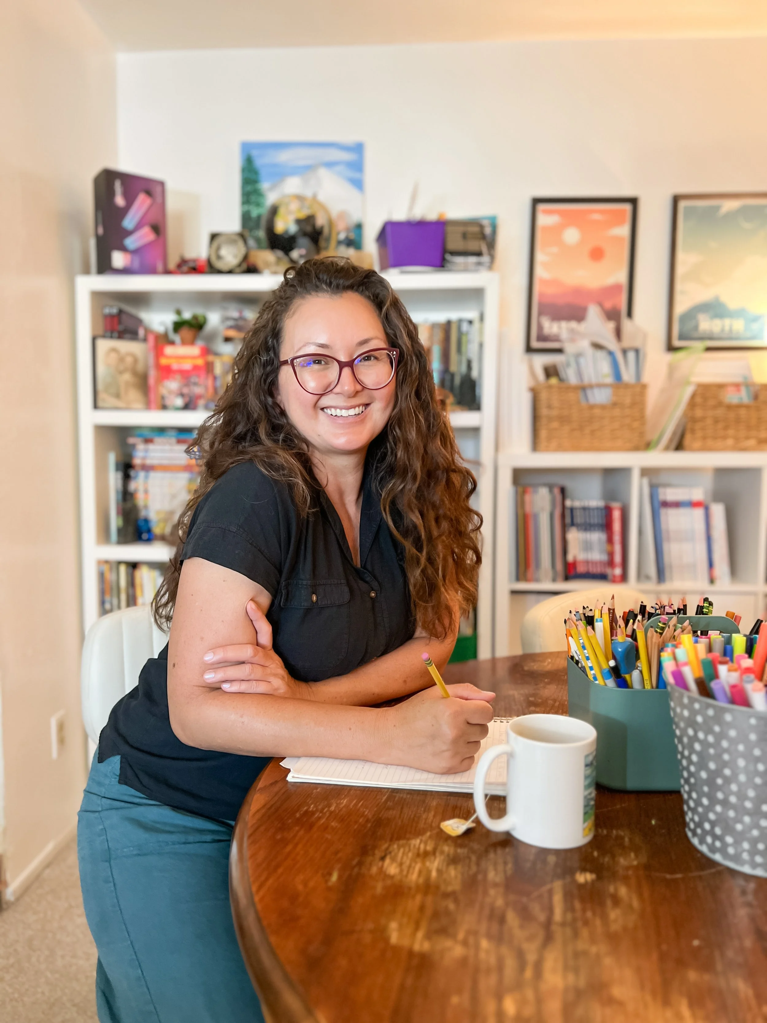 A woman with long curly hair and glasses smiling at the camera, sitting at a wooden table with a notebook and colored pencils, in a room with bookshelves and framed posters on the wall.