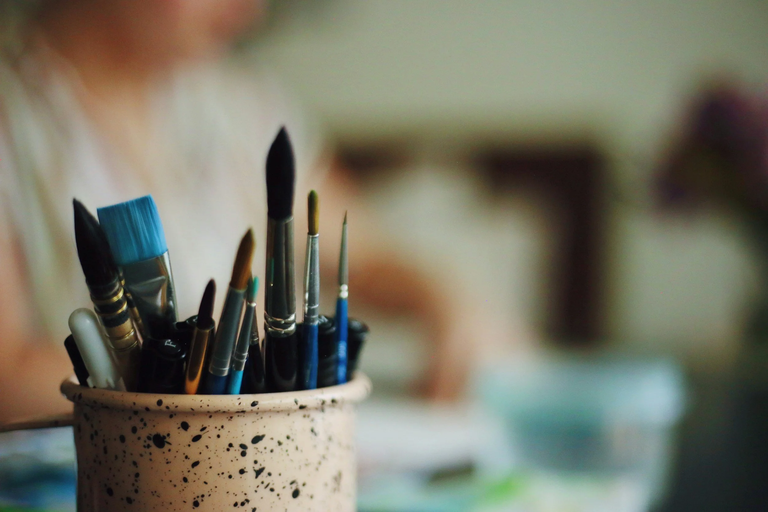 A white speckled ceramic mug holding a variety of paintbrushes with different sizes and shapes, set against a blurred colorful background.