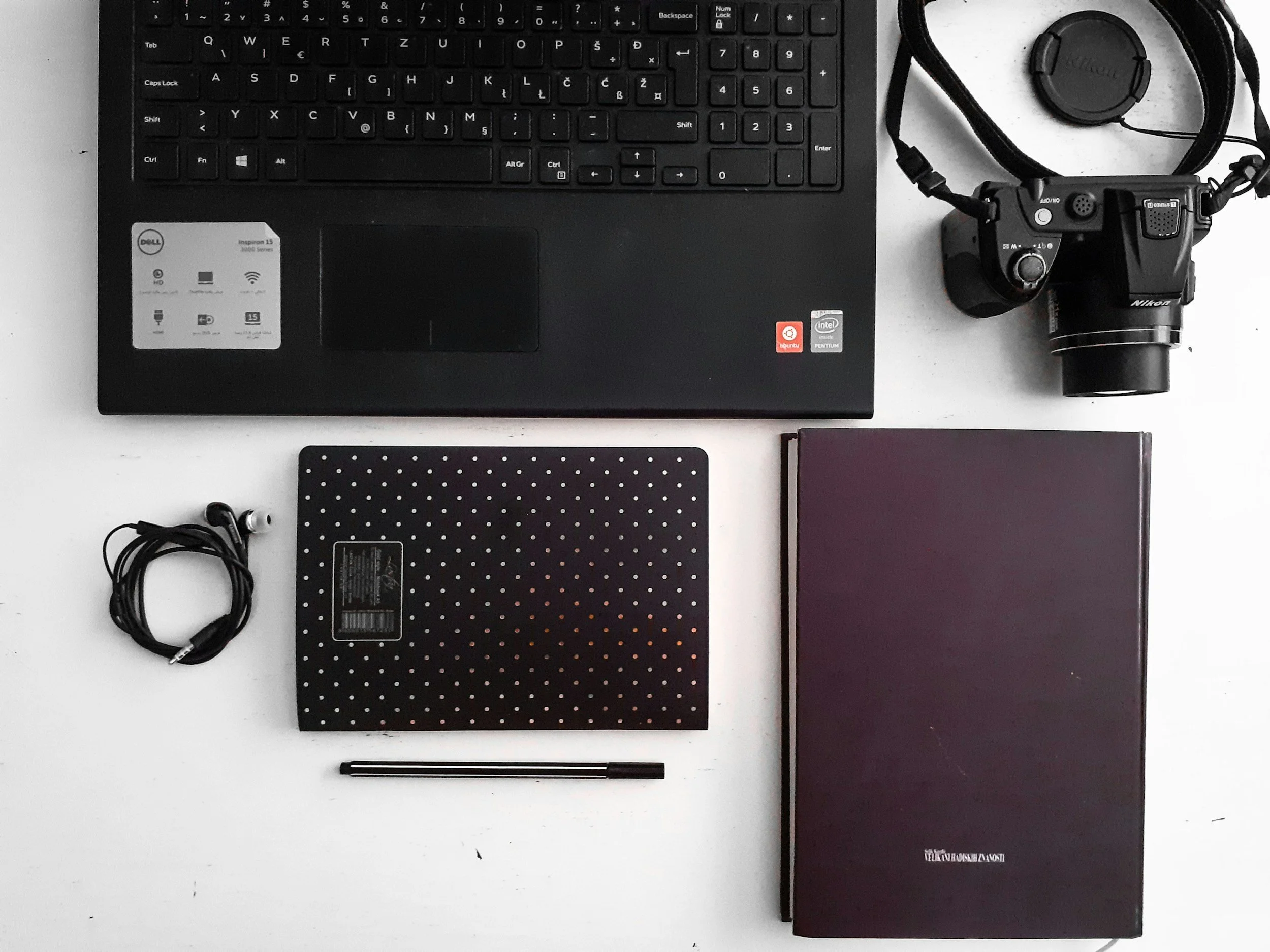 Top view of a workspace with a black keyboard, a wireless mouse, a camera, a closed notebook, a pen, a pair of earphones, and a dotted notebook on a white surface.