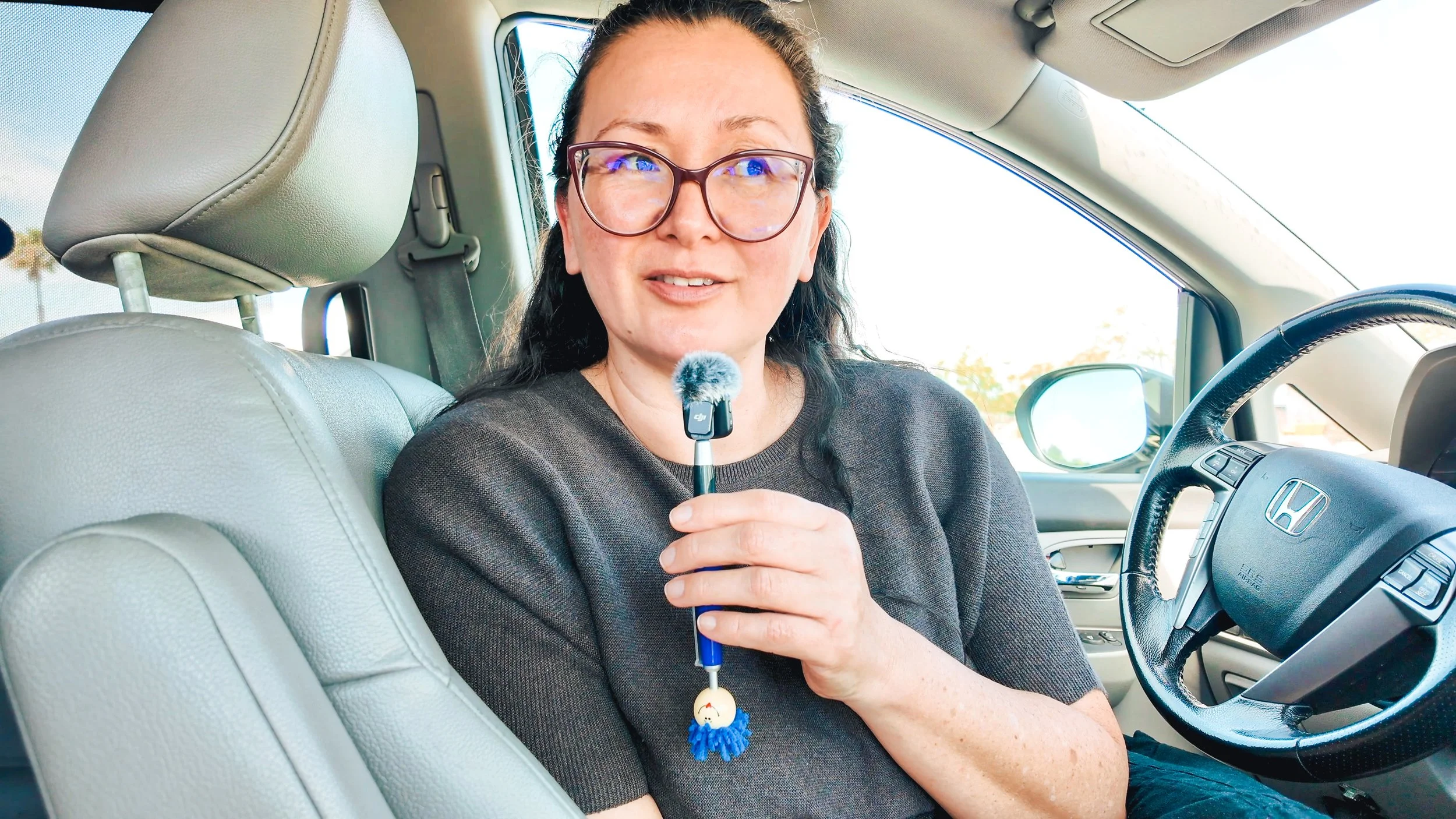 A woman sitting in the driver's seat of a car, holding a small handheld microphone with a fuzzy windscreen, wearing glasses and a black shirt, with a beige interior and a Honda steering wheel visible.