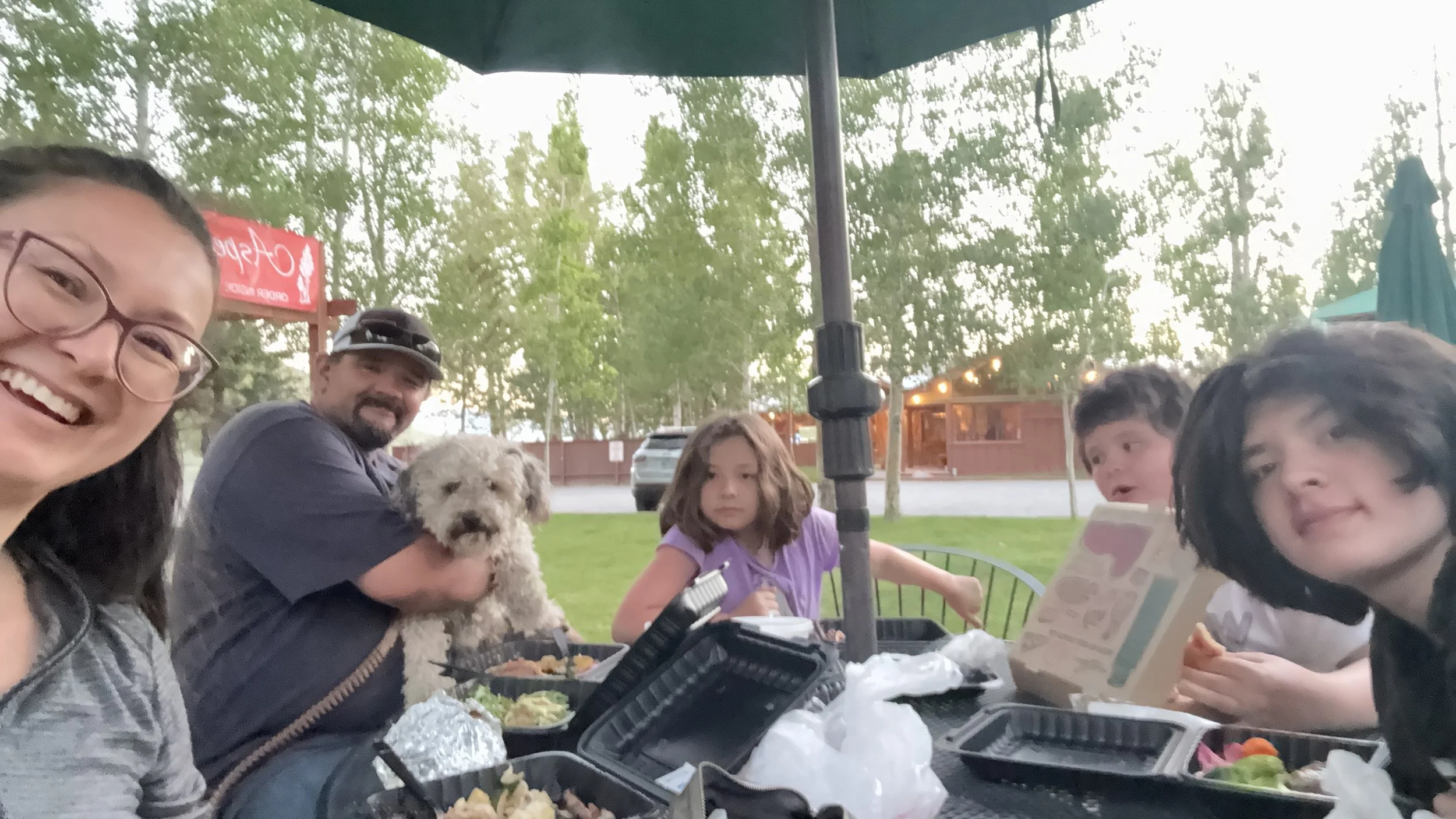A group of people, including two children and adults, sitting outdoors at a table with food and drinks under a green umbrella. One person is holding a small fluffy dog, and others are eating or looking at menus. Trees and a fence are visible in the background.