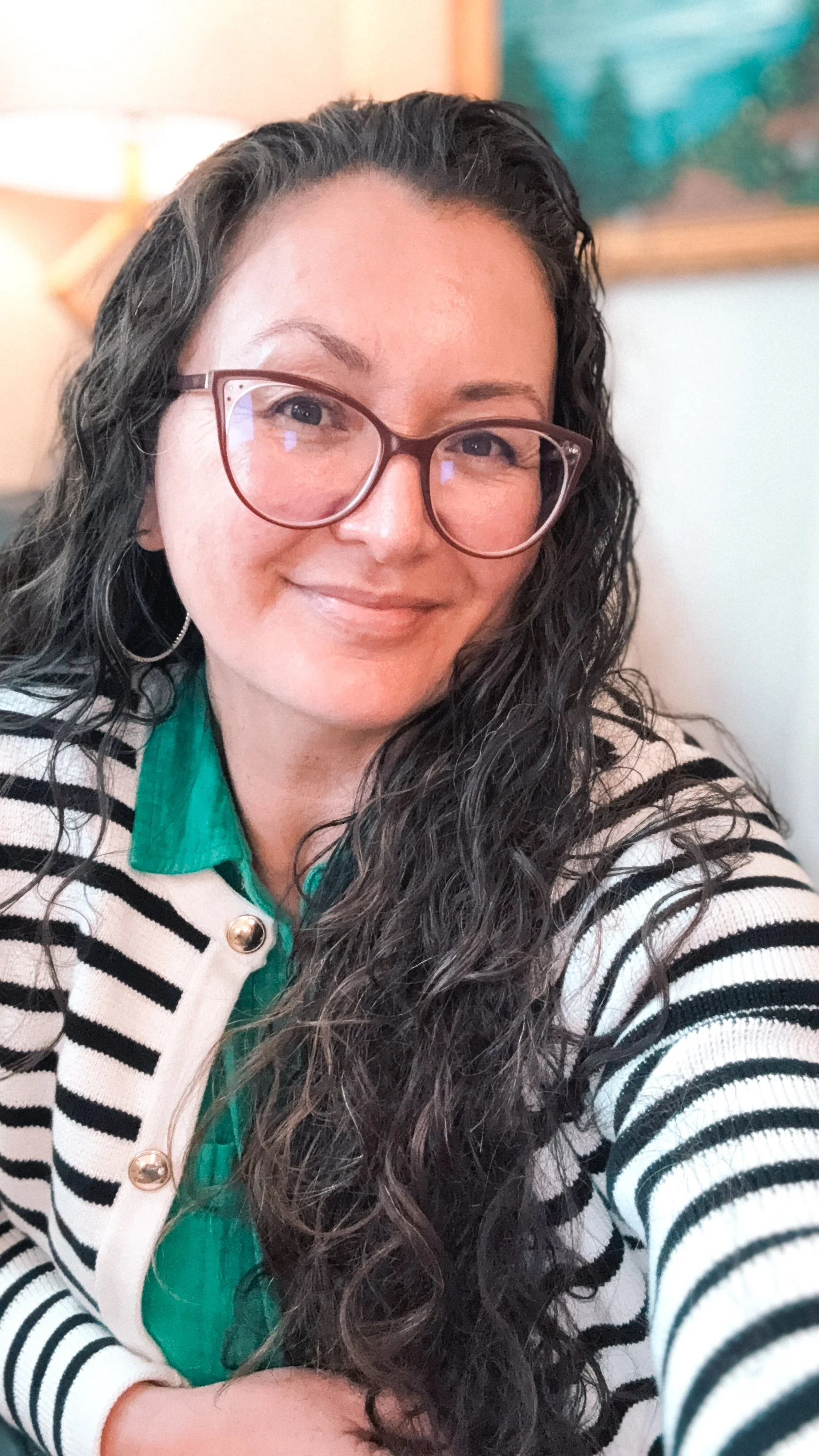 A woman with long dark curly hair wearing glasses, a green shirt, and a black and white striped cardigan smiling at the camera.