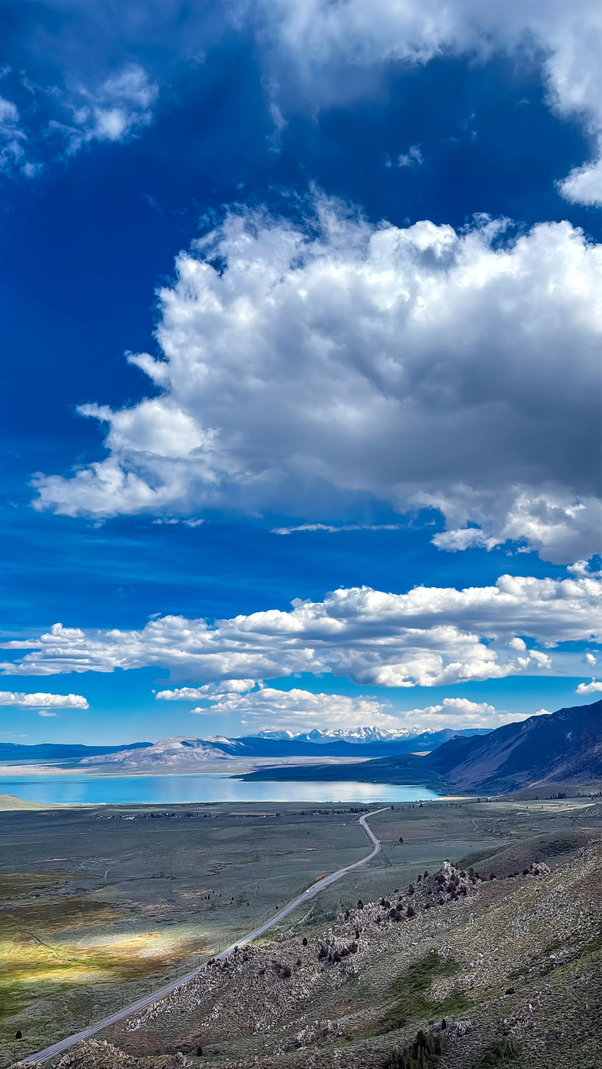 Scenic landscape with a vast lake, mountains in the distance, green rolling hills, and a partly cloudy sky.