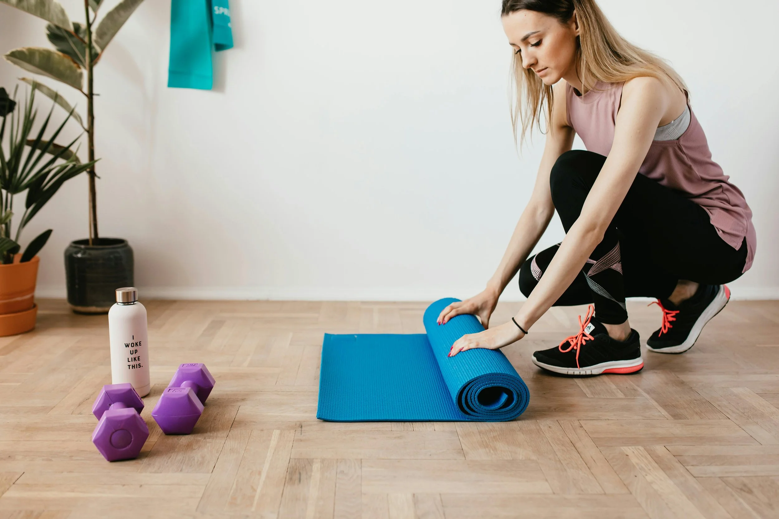 A woman is rolling out a blue yoga mat in a home gym, with purple dumbbells, a water bottle, and a potted plant nearby.