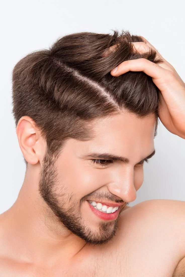 Close-up of a smiling man with dark brown hair and a well-groomed beard, touching his hair with his right hand.