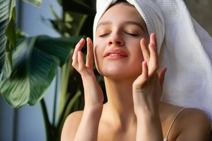 A woman with a towel wrapped around her head, eyes closed, gently touching her face, possibly in a spa or skincare setting, with green tropical plants in the background.