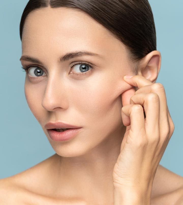 A woman with fair skin and blue eyes touching her face, with her hand close to her cheek, against a light blue background.