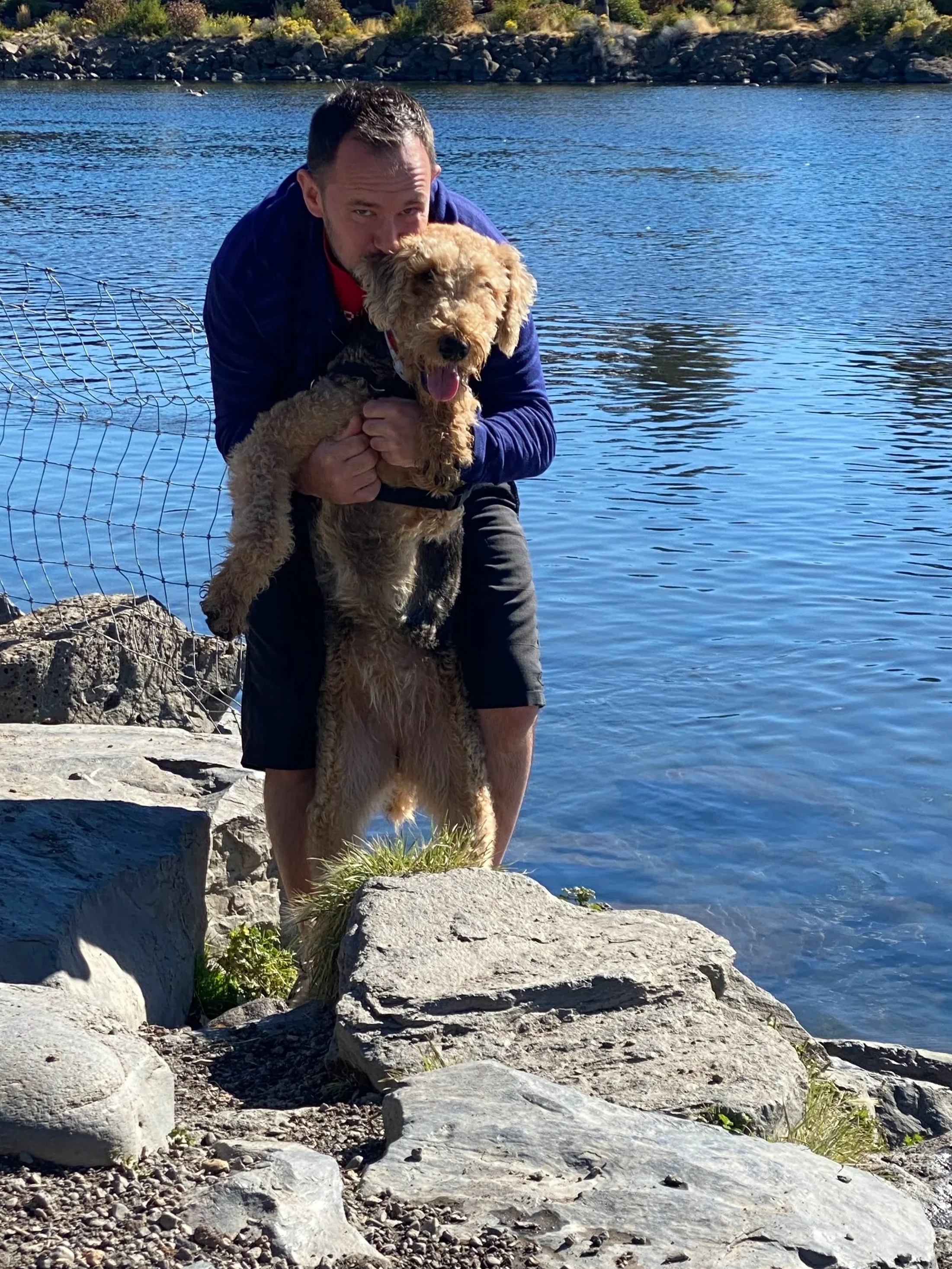 Picture of man hugging Airedale terrier dog on a rocky riverfront