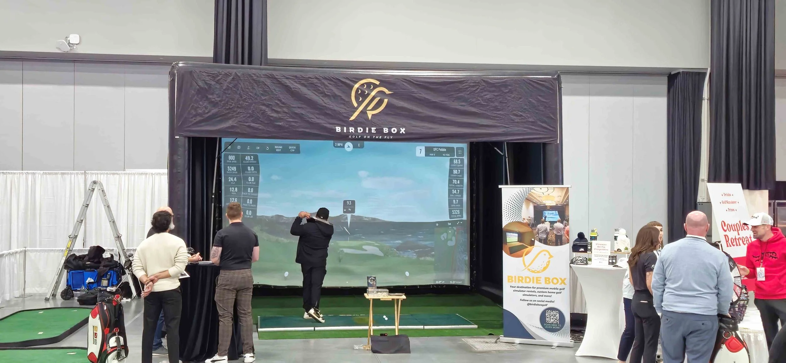 Indoor golf simulation event with multiple people, a large screen displaying a golf course, and a 'Birdie Box' booth promoting premium mobile golf simulations.