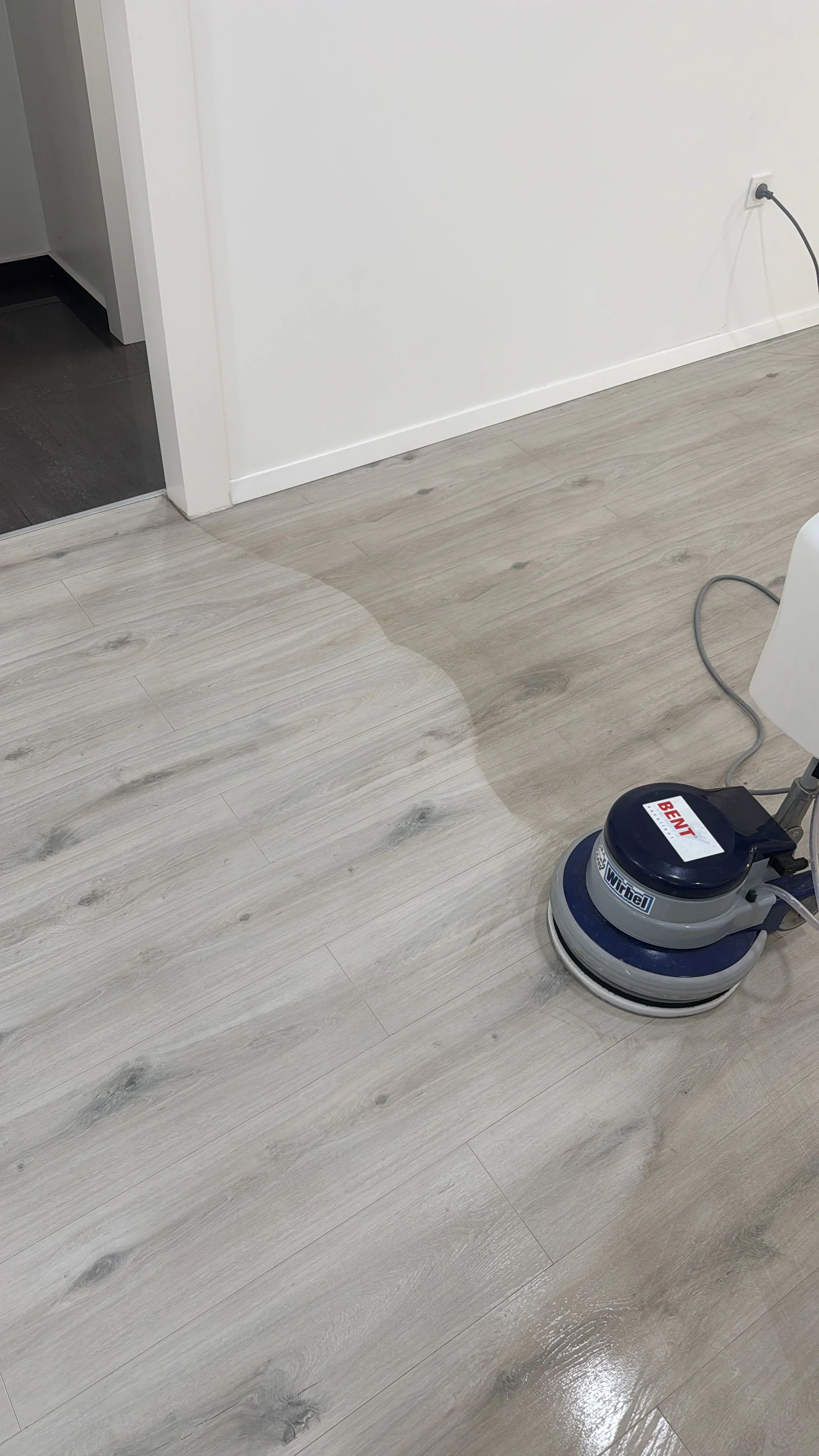 A floor cleaning machine on light wood flooring near a white wall and electrical outlet.