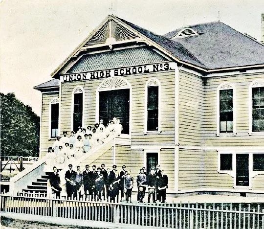 Black and white historic photograph of students and teachers standing in front of Union High School No. 3, a wooden building with a sign on top, stairs leading to the entrance, and a fence in the foreground.