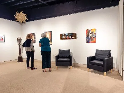Two women looking at artwork in an art gallery with three paintings on the wall, a sculpture on a pedestal, and two black armchairs.