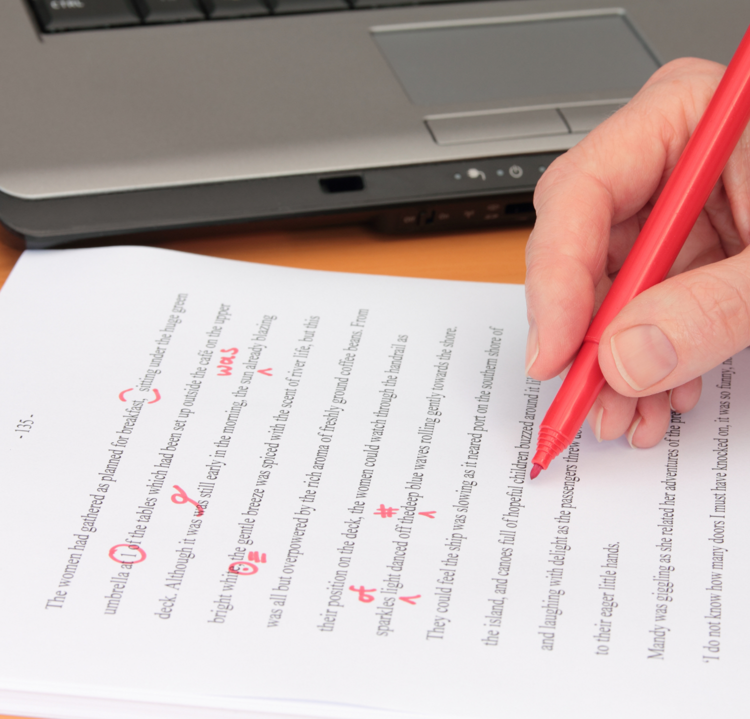 Close-up of a hand holding a red pen, marking edits on a printed document with red ink, in front of a laptop computer.