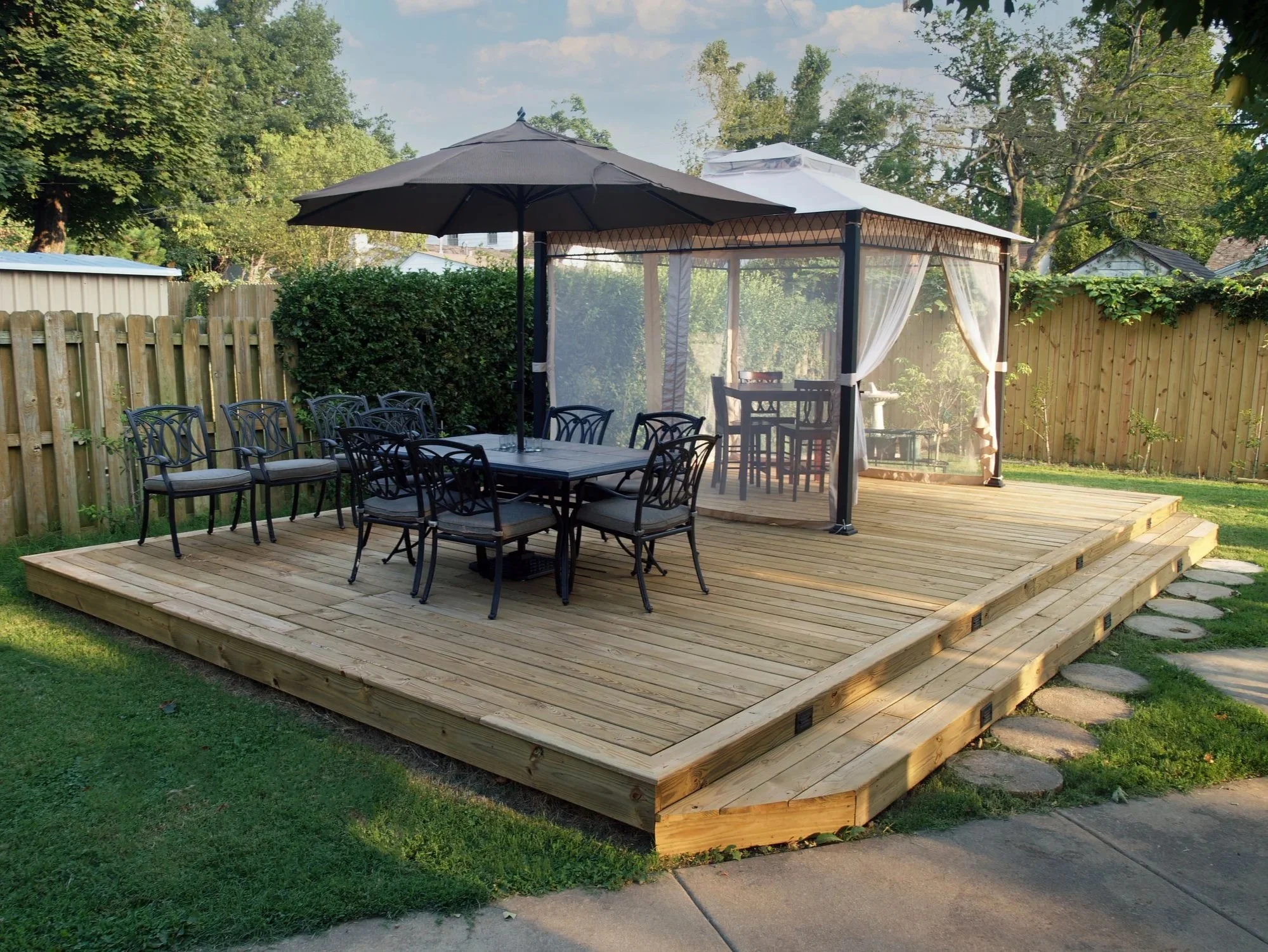 A backyard wooden deck with outdoor dining furniture, a large black patio umbrella, and a gazebo with curtains, surrounded by a wooden fence and lush greenery.