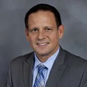 Portrait of a man in a gray suit and blue tie, smiling against a gray background.