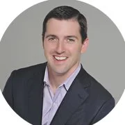 Professional headshot of a smiling man in a suit and light shirt against a gray background.