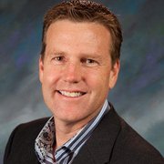 Professional headshot of a smiling man with short, light brown hair, wearing a dark blazer over a collared shirt with a patterned inner shirt.