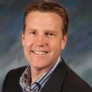 A portrait of a smiling man with short brown hair, wearing a dark blazer and light collared shirt, against a blurred background.