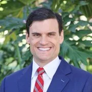 A man in a blue suit, white shirt, and red striped tie smiling outdoors with green leafy background.