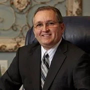 A man in a suit and tie sitting at a desk in an office setting, smiling at the camera.