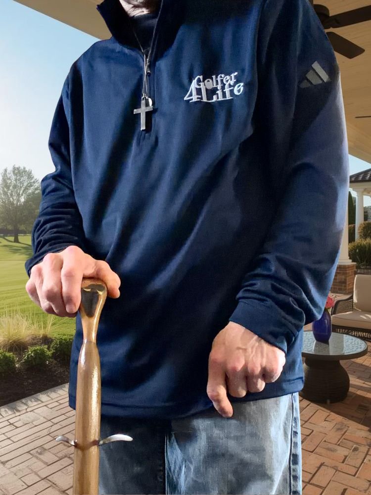 A person wearing a dark blue jacket with 'Golfer 4 Life' embroidered on it, holding a golf club, standing on a patio with outdoor furniture and a garden background.